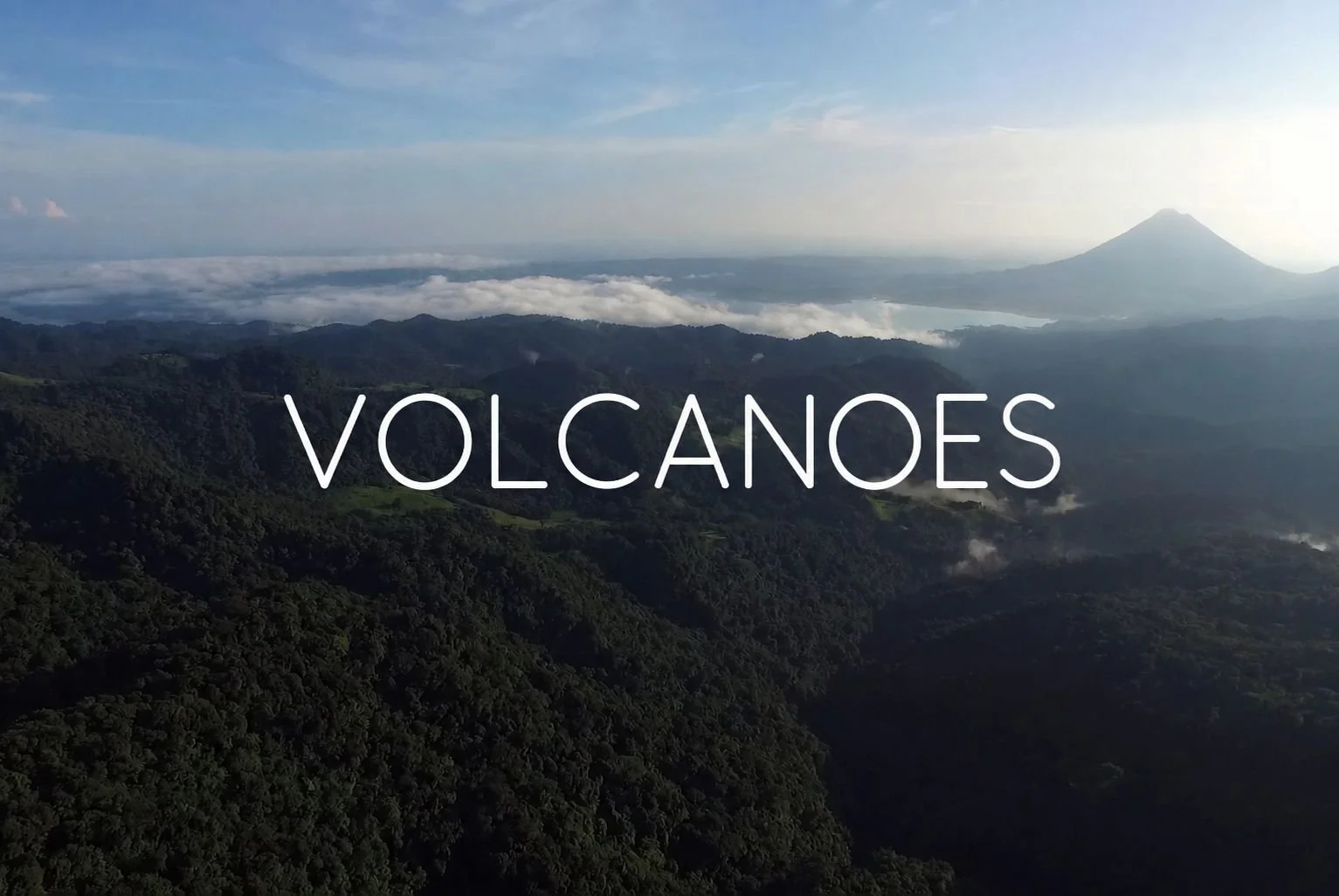 Aerial view of lush green mountains with a volcano in the distance and clouds hovering over the landscape, with the word 'VOLCANOES' overlayed.