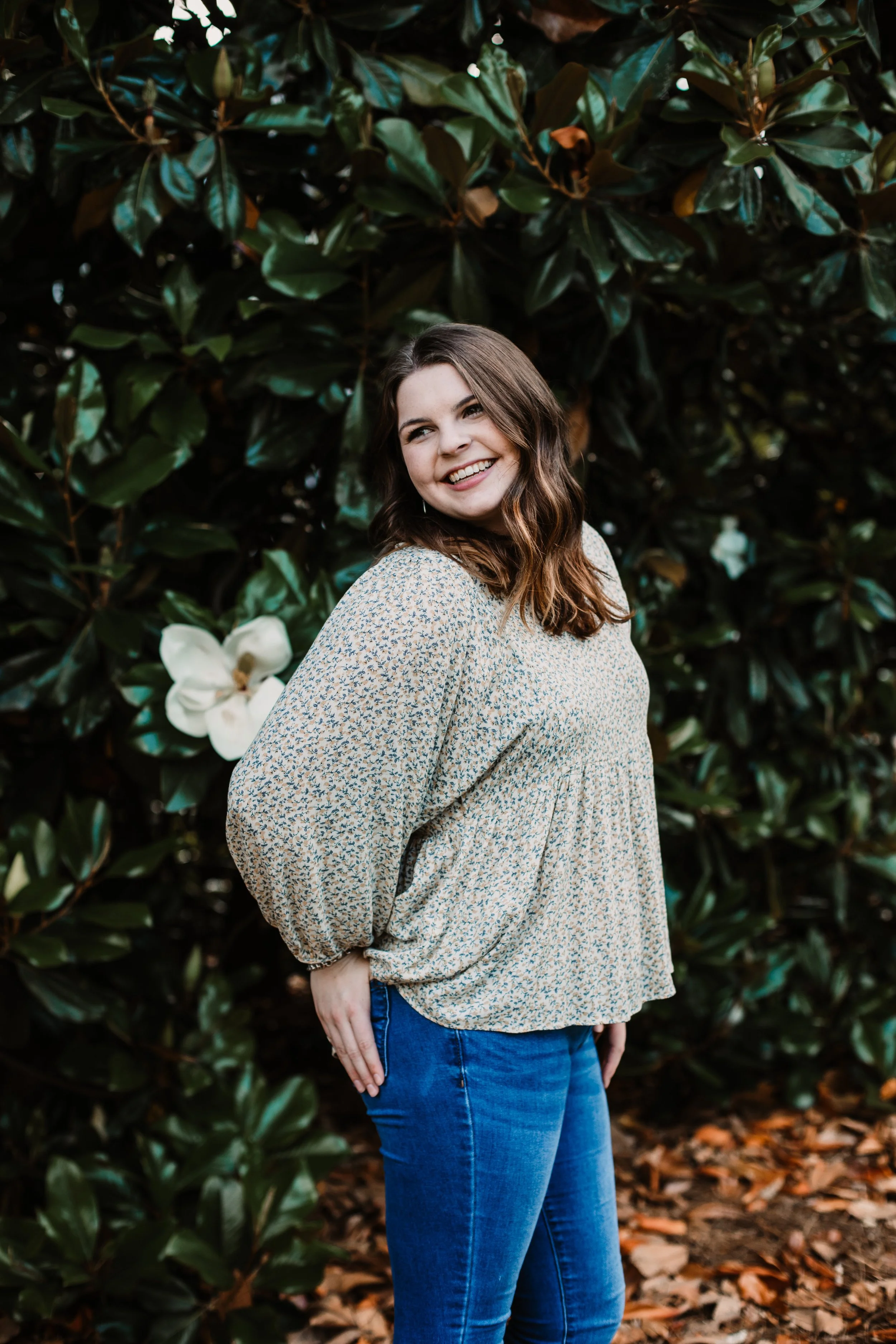 Smiling woman in a floral blouse and blue jeans standing among lush green foliage and white flowers