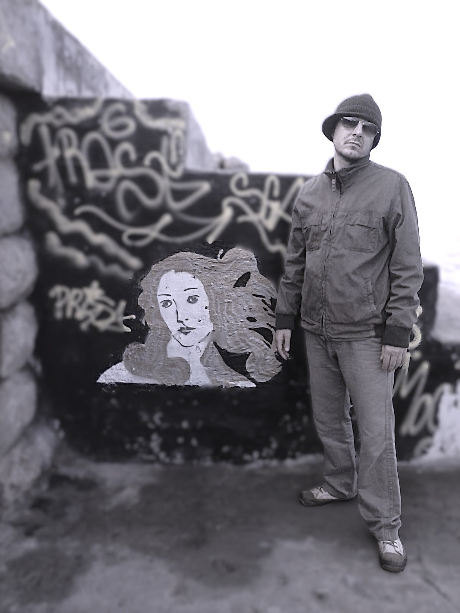 A man wearing a beanie, glasses, a gray jacket, and gray pants standing outdoors near graffiti-covered rocks with a painted woman's face on one of the rocks.