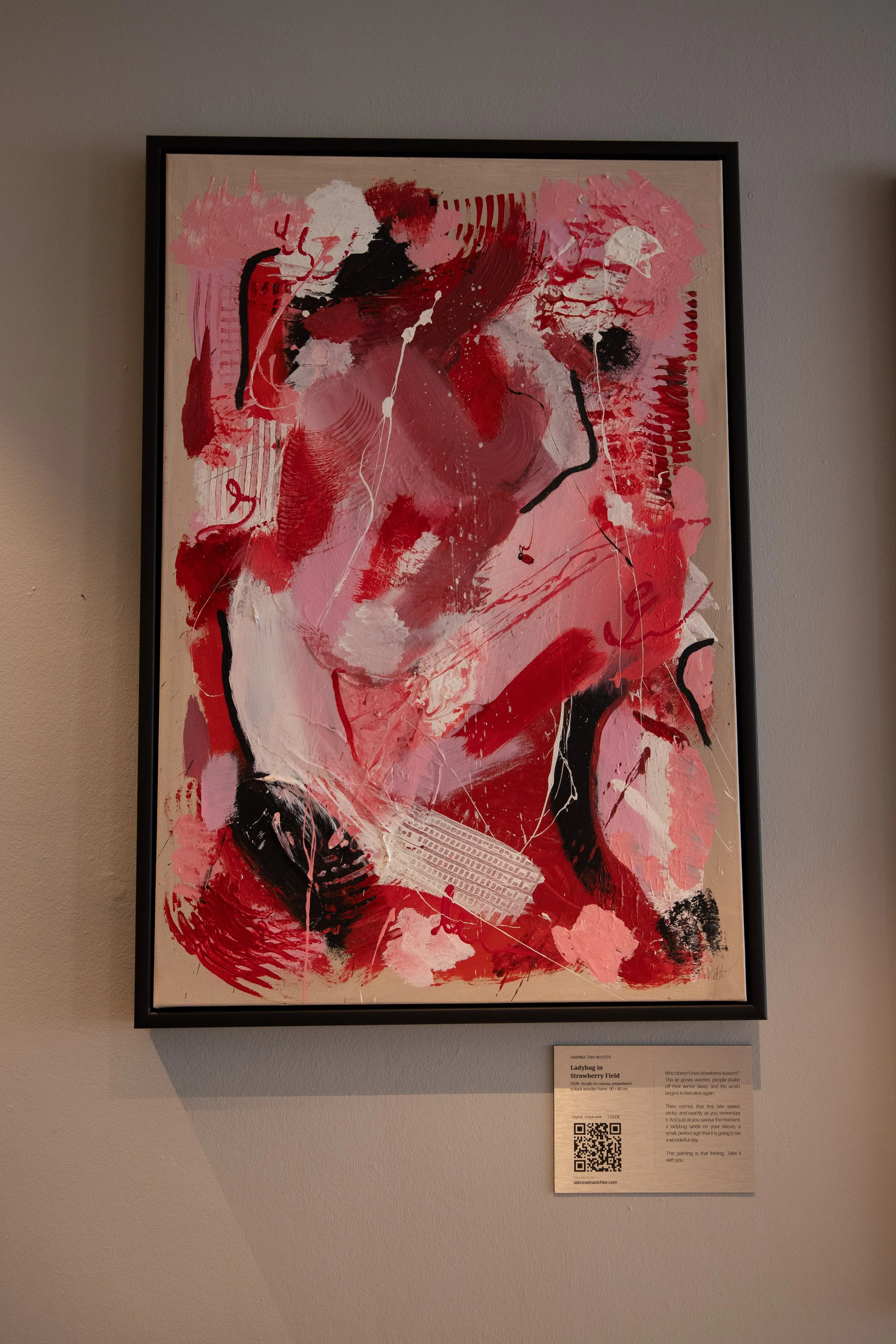 Abstract painting with red, pink, black, and white brushstrokes, textures, and lines in a black frame on a gallery wall.