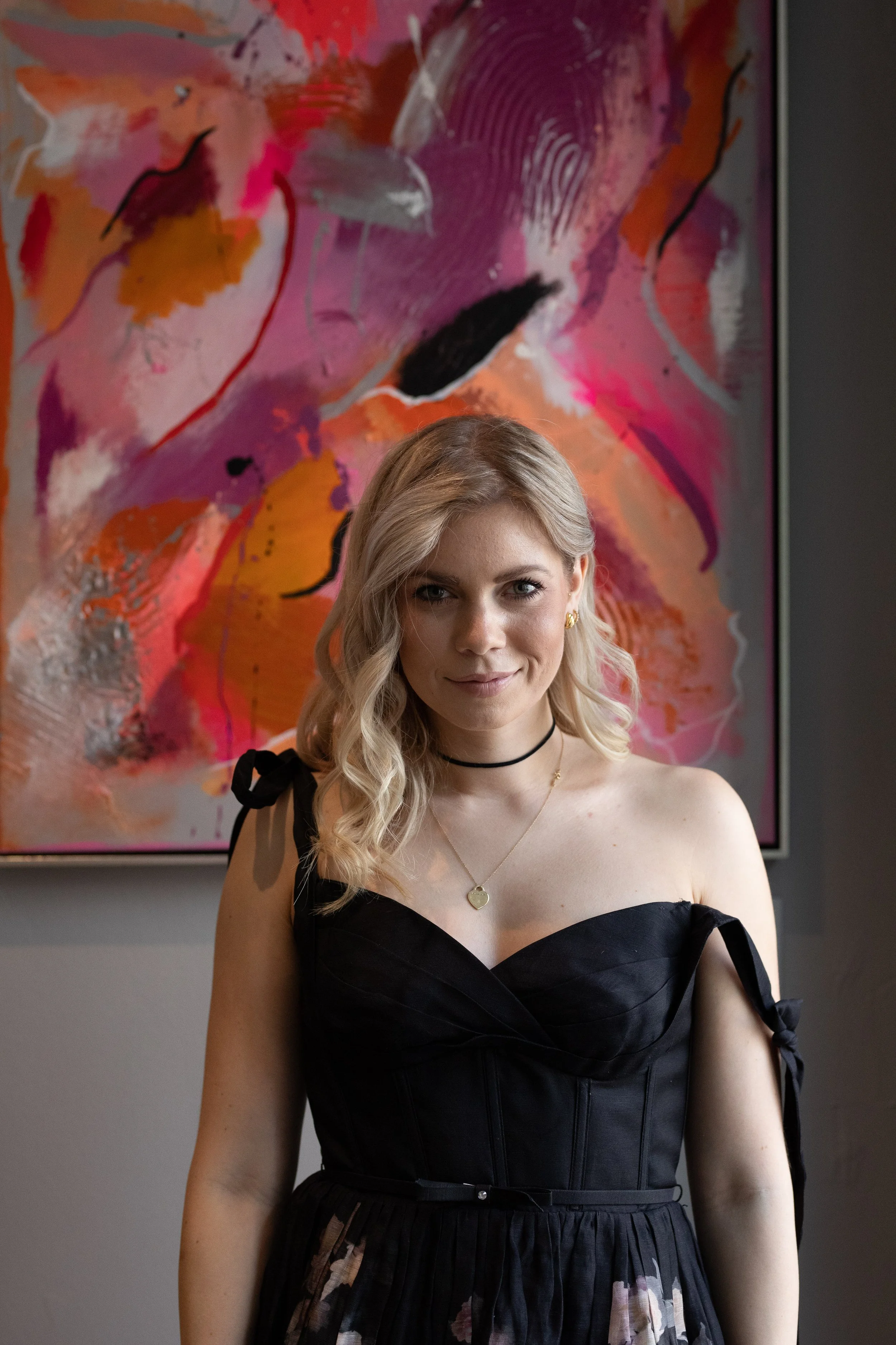 Young woman with blonde hair and blue eyes wearing a black dress with puffed sleeves and a pleated skirt, standing in front of an abstract floral painting with shades of pink, orange, purple, and black.