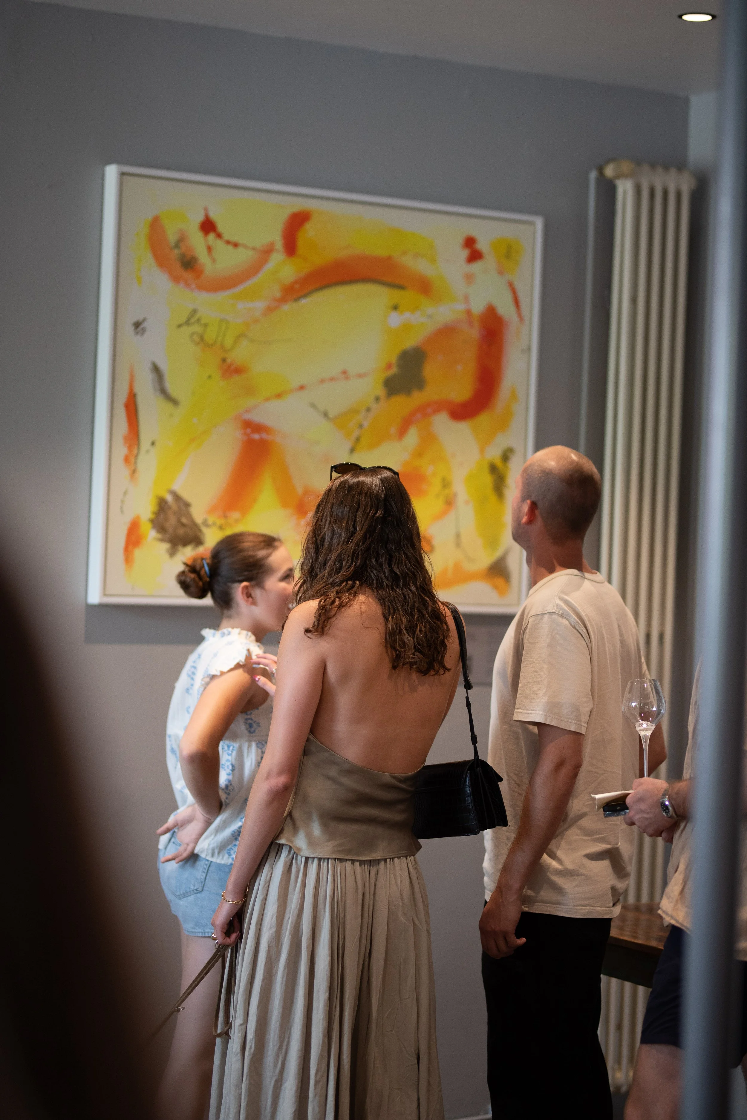 Group of four people standing in front of colorful abstract painting in an indoor art gallery. Two women and two men are engaged in conversation, with a wine glass held by one of the individuals.