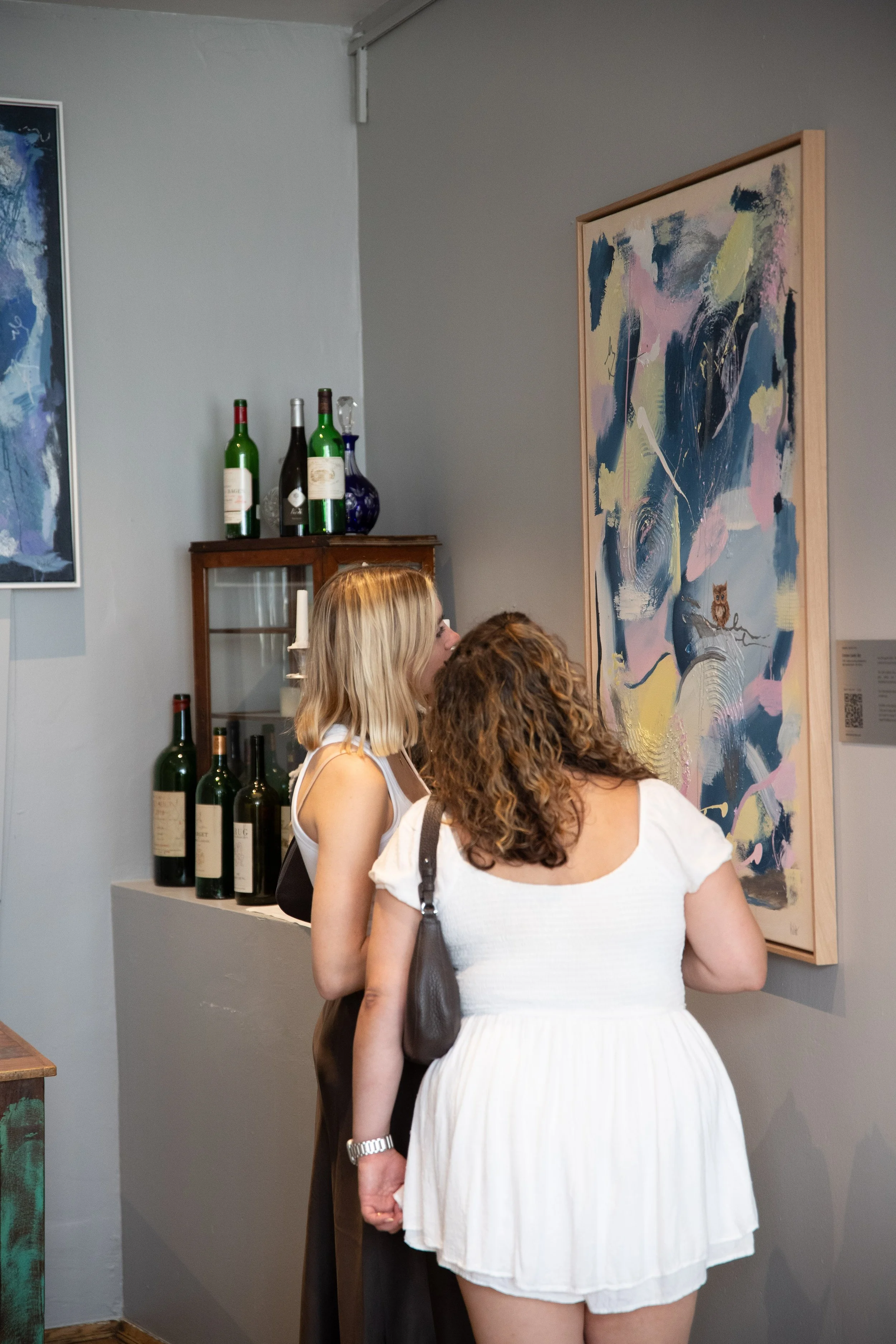 Three women browsing artwork in an art gallery, with paintings on the wall and bottles on display in the background.