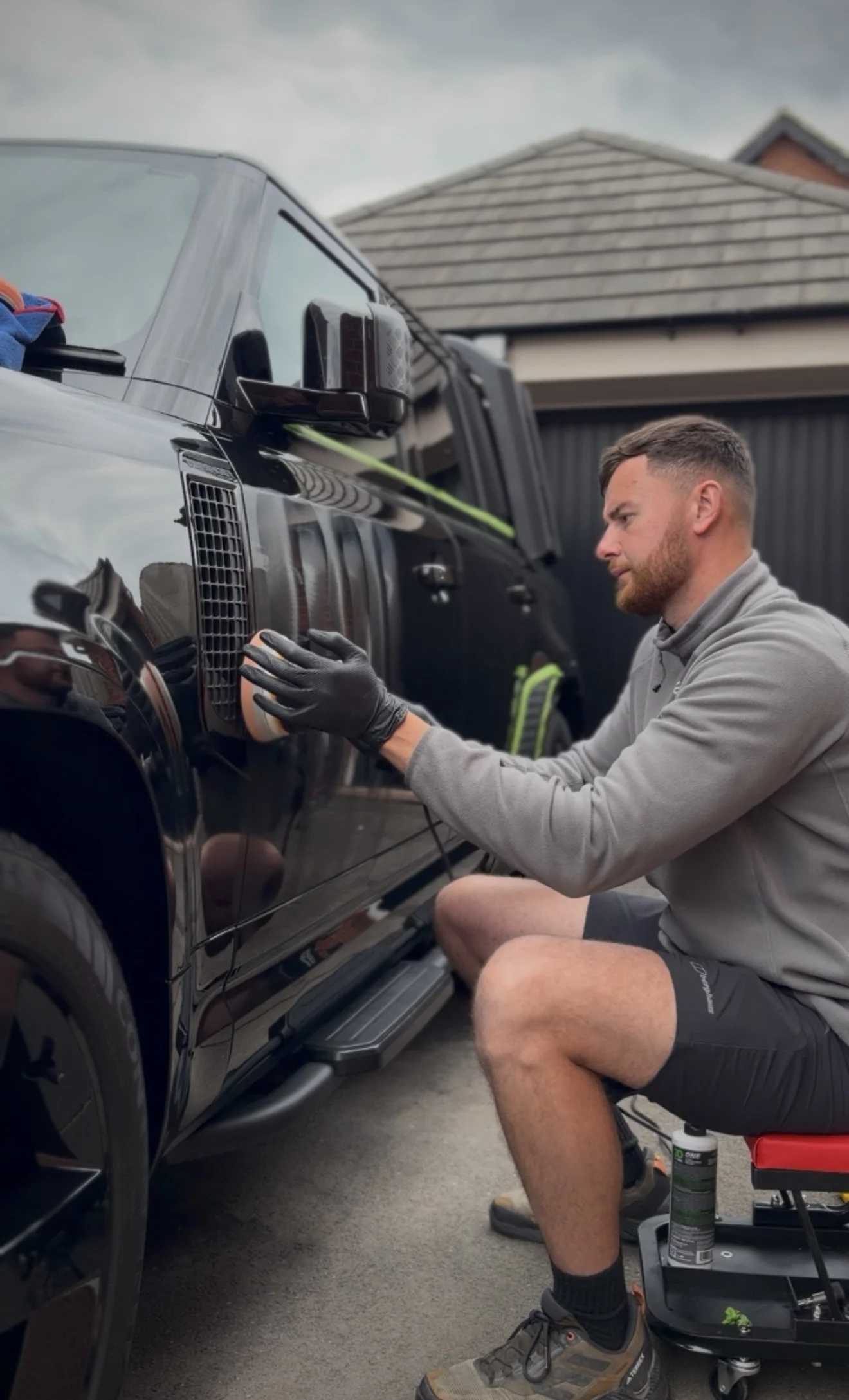 A man with short hair, a beard, and wearing a gray hoodie and shorts, is kneeling on a rolling seat cleaning the side of a black SUV with a polishing pad. There is a house with a tiled roof in the background, and the sky appears cloudy.