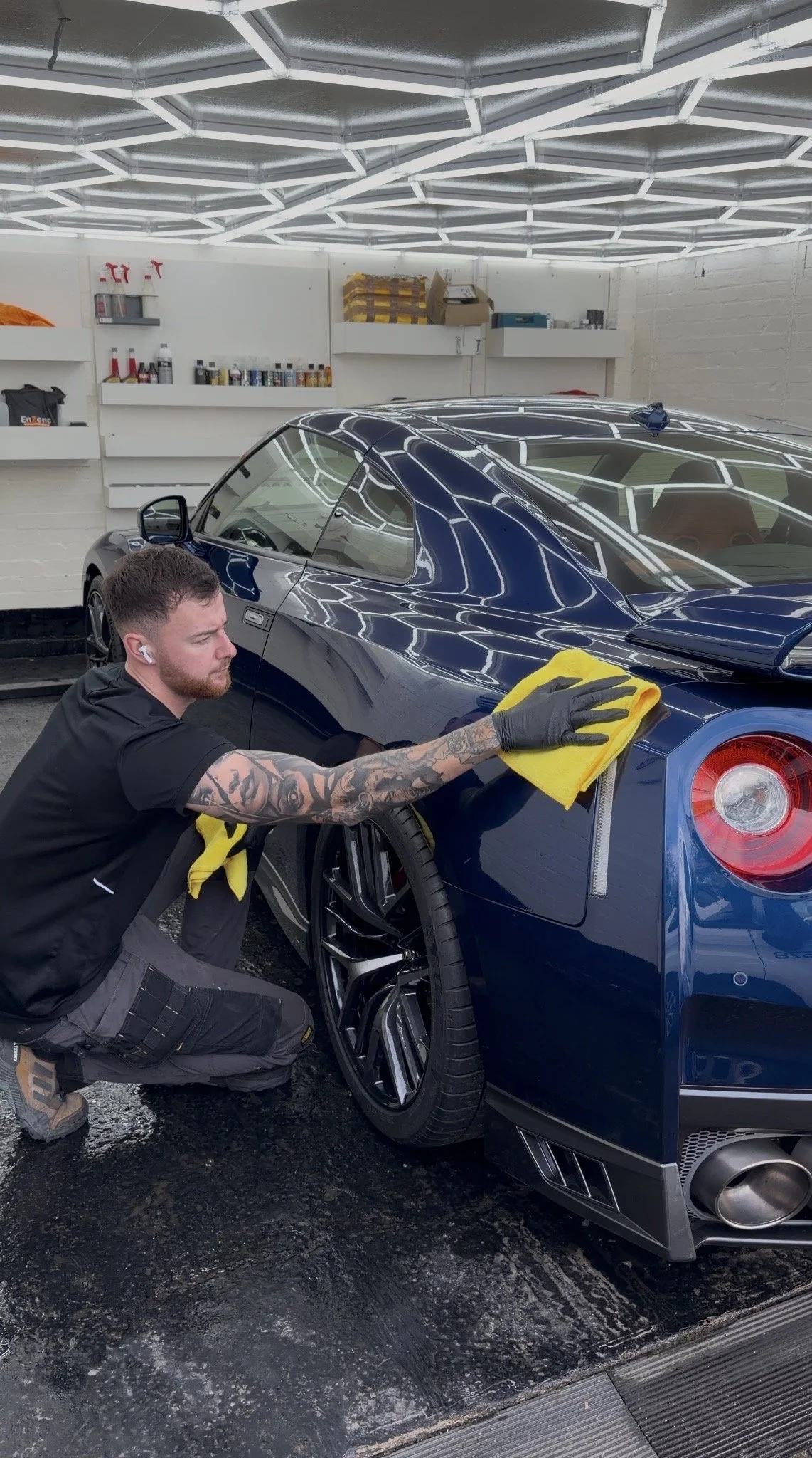 A man is polishing a blue sports car inside a garage, using a yellow cloth. The garage has white walls, shelves with various supplies, and an intricate ceiling light design.