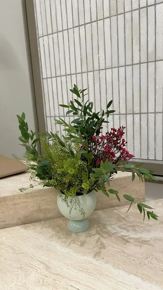 A white ceramic vase contains a mixed bouquet of green foliage and pink flowers, placed on a beige marble surface near a window with a striped wall in the background.