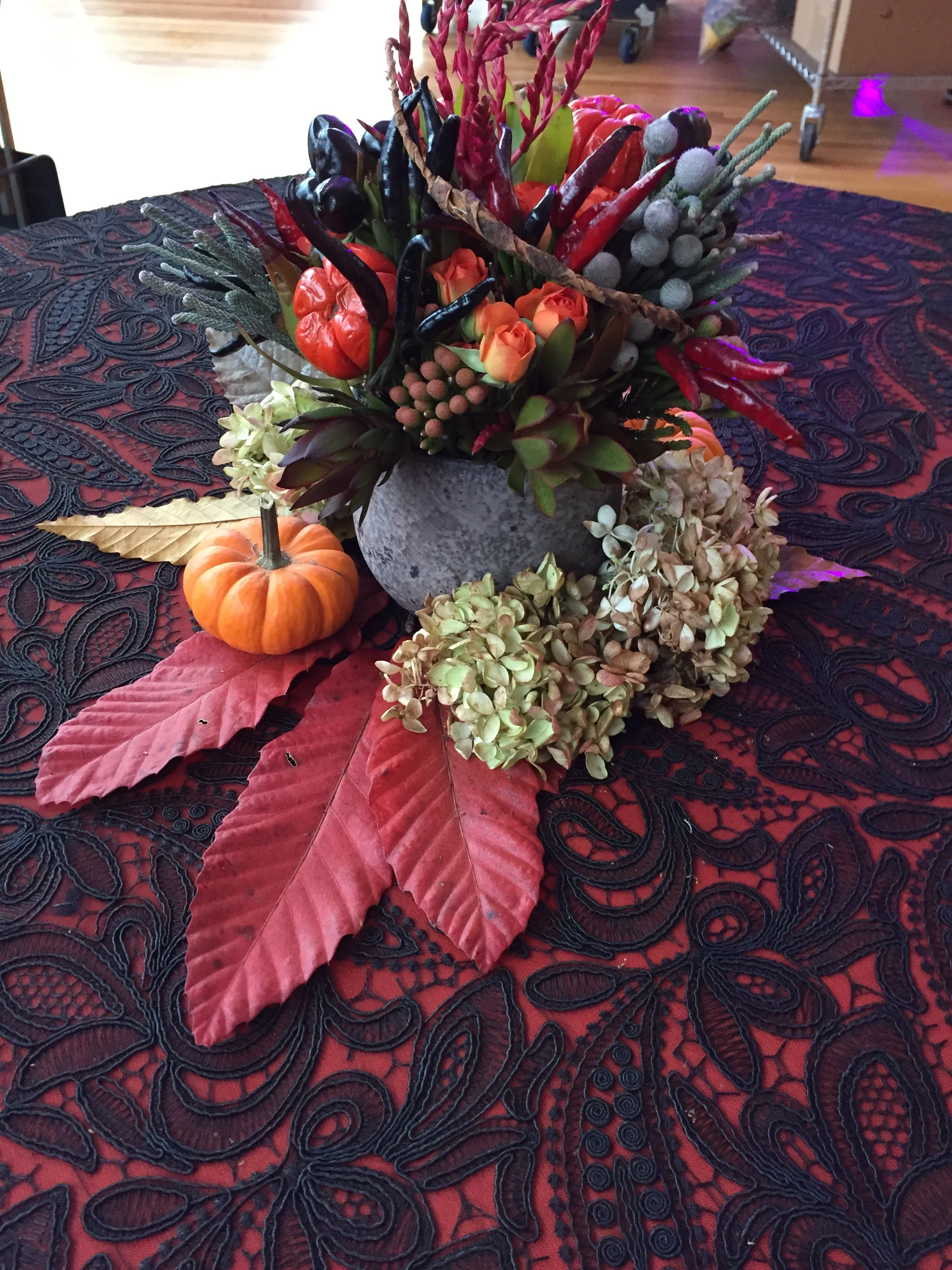 A table centerpiece with artificial pumpkins, red chili peppers, ornamental berries, and leaves on a textured red and black tablecloth.
