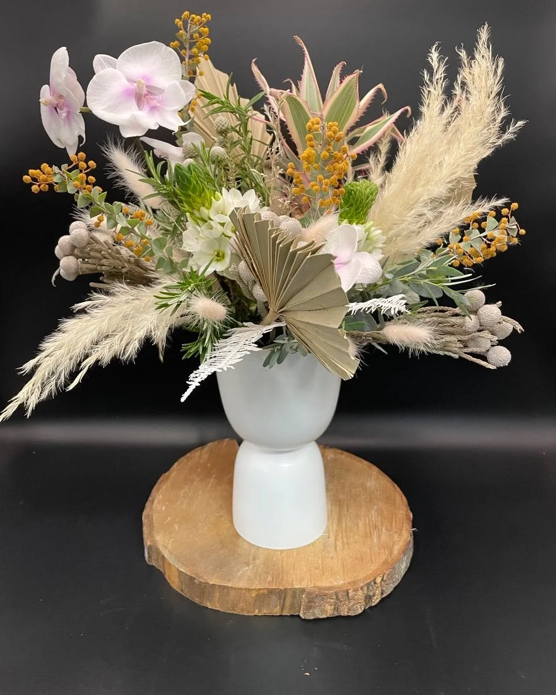 A bouquet of artificial flowers with white, pink, and yellow accents arranged in a white vase, placed on a wooden slab against a dark background.