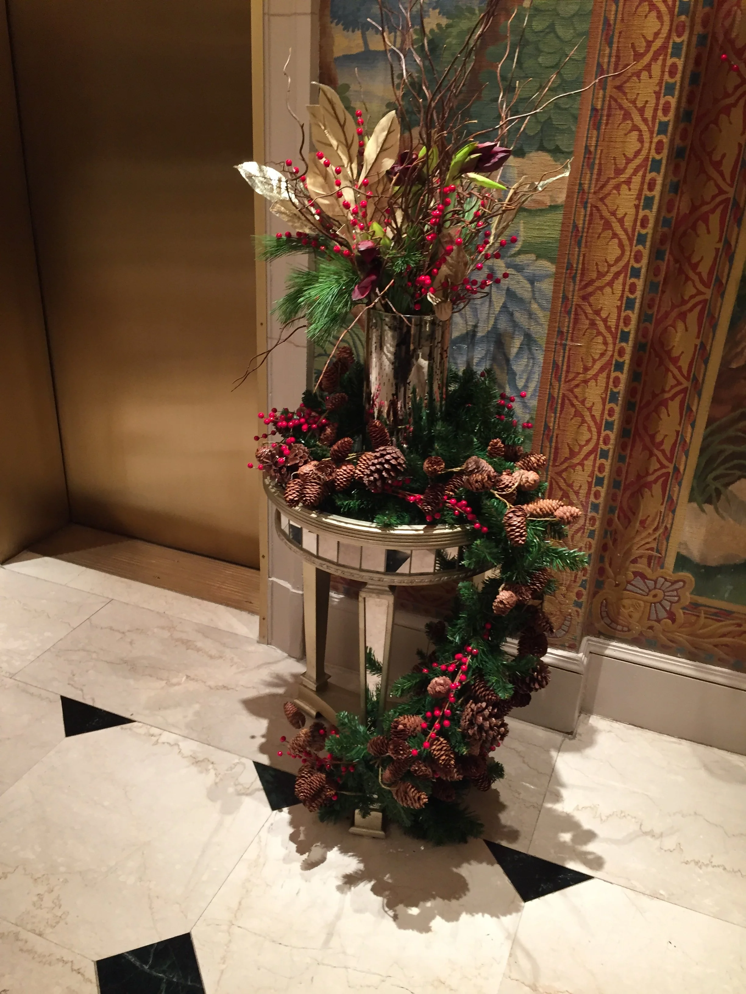 A tall decorative floral arrangement with pinecones, red berries, branches, and leaves in a silver vase, placed on a small round table with a marble floor and a tapestry hanging on the wall behind.