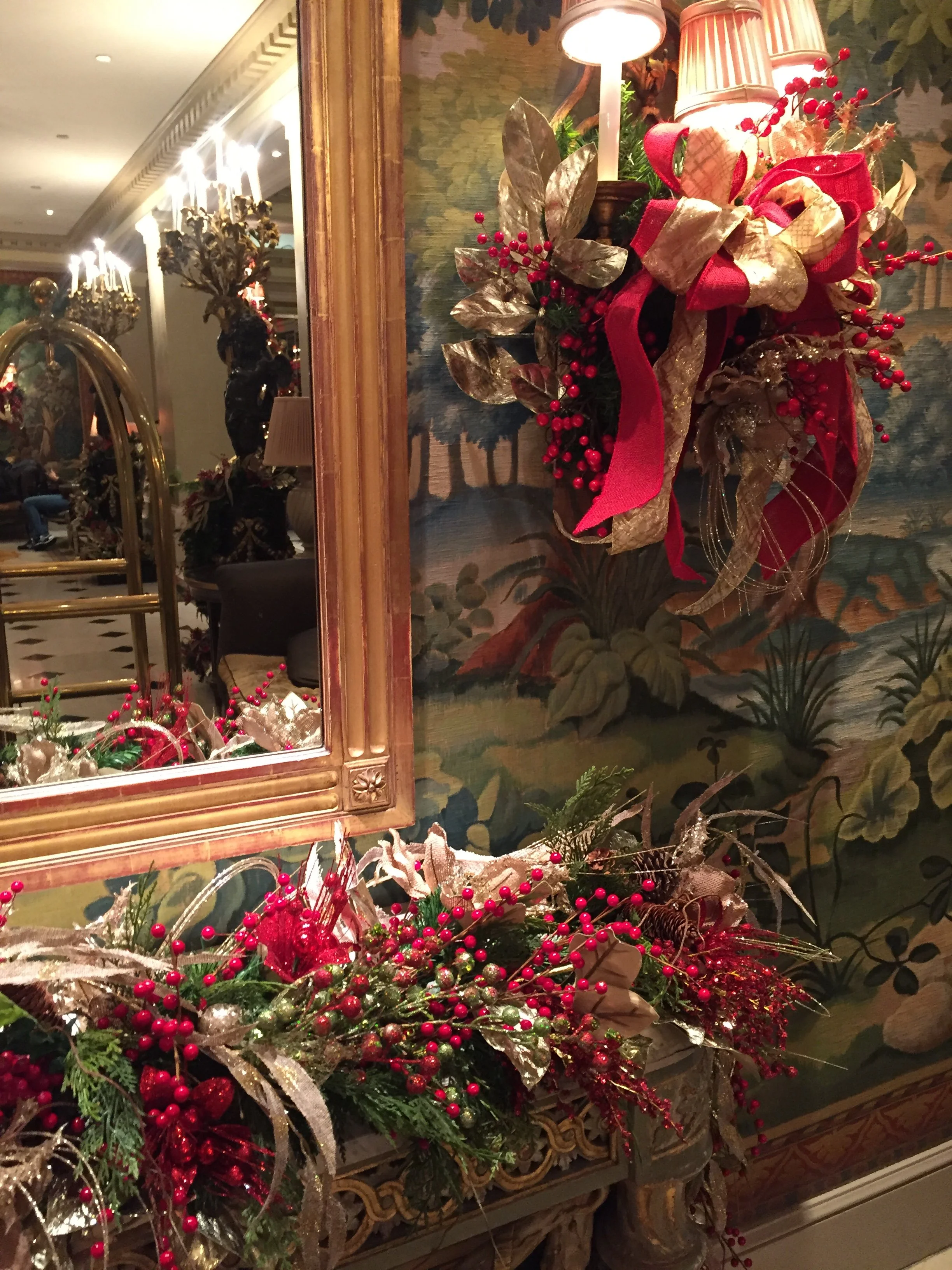 Festive holiday floral arrangement with red berries, gold and red ribbons, and decorative leaves in a vase, in front of a mirror and wall with paintings in a decorated interior space.