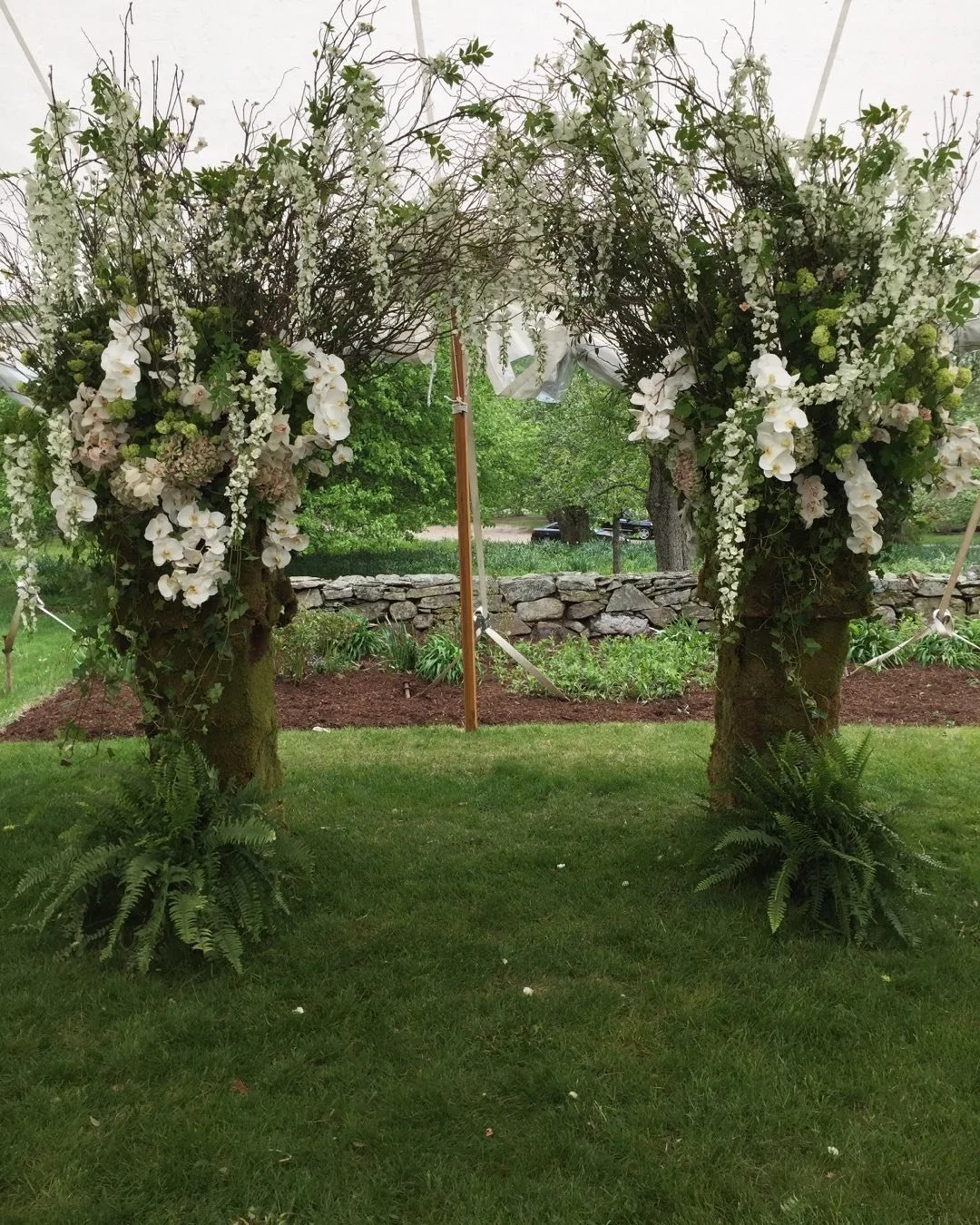 A floral arch with white flowers and greenery on moss-covered tree trunks, set up outdoors on a grassy area with a stone wall and trees in the background.