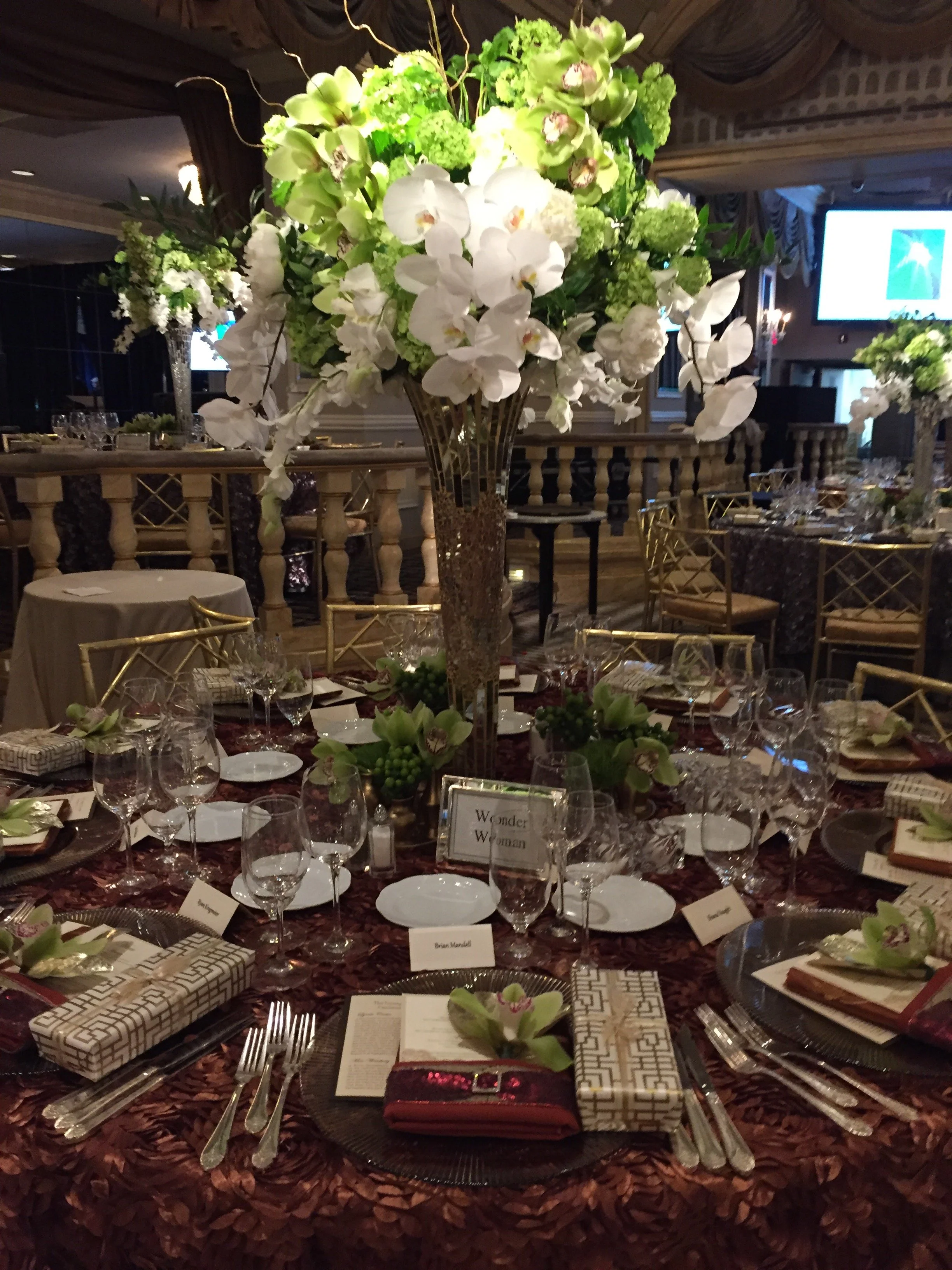 Elegantly set banquet table with a tall floral centerpiece of white orchids and green hydrangeas. The table is decorated with patterned napkins, place cards, glassware, and silverware, in a luxurious setting with additional floral arrangements in the