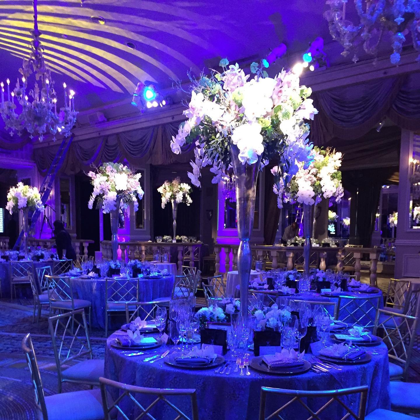 Elegant event space decorated with tall flower arrangements on tables, chandeliers, and purple lighting.