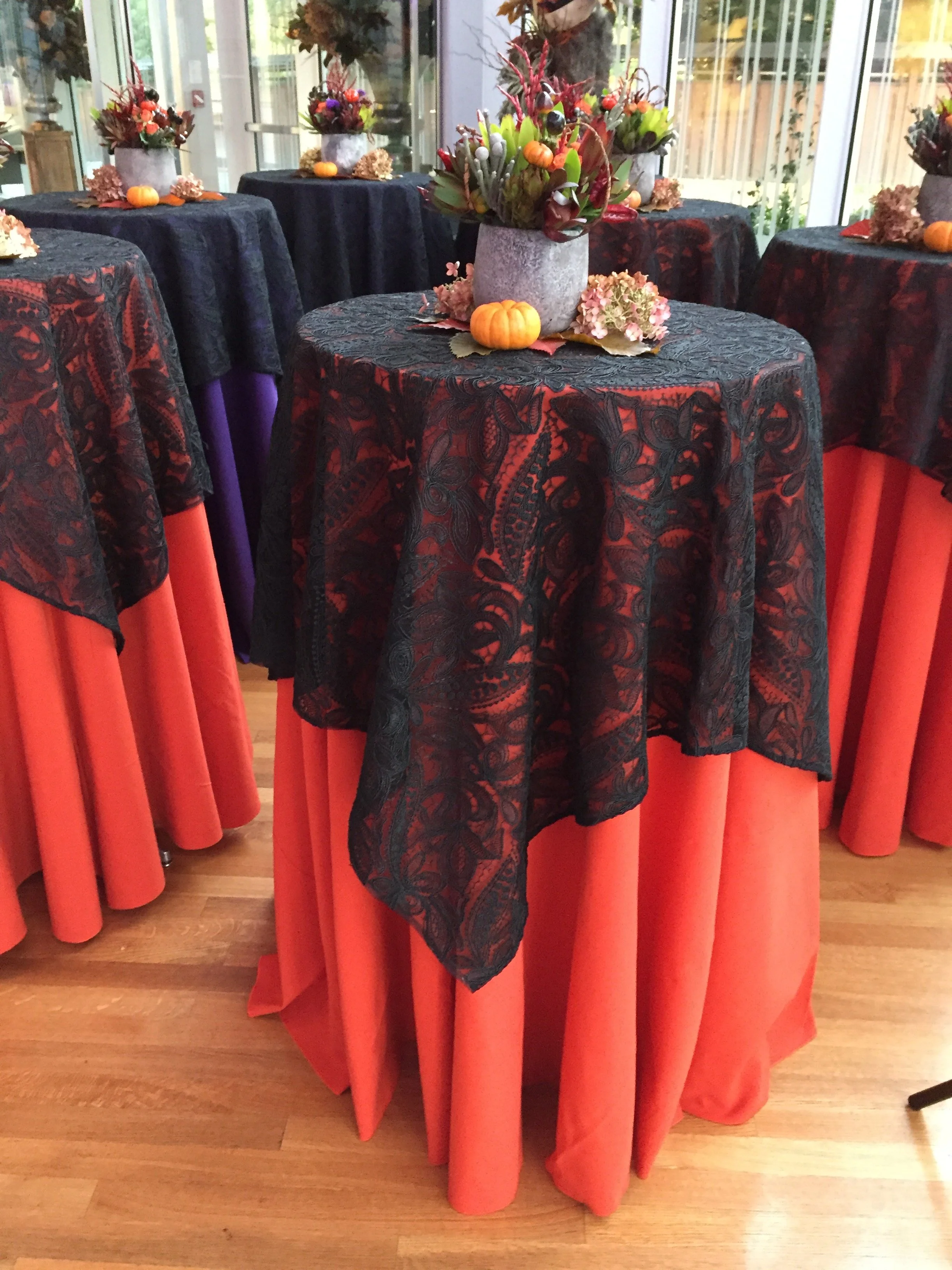A round table decorated with autumn-themed floral arrangements, small pumpkins, and fall leaves, covered with black lace tablecloth, inside a room with large windows and wooden flooring.