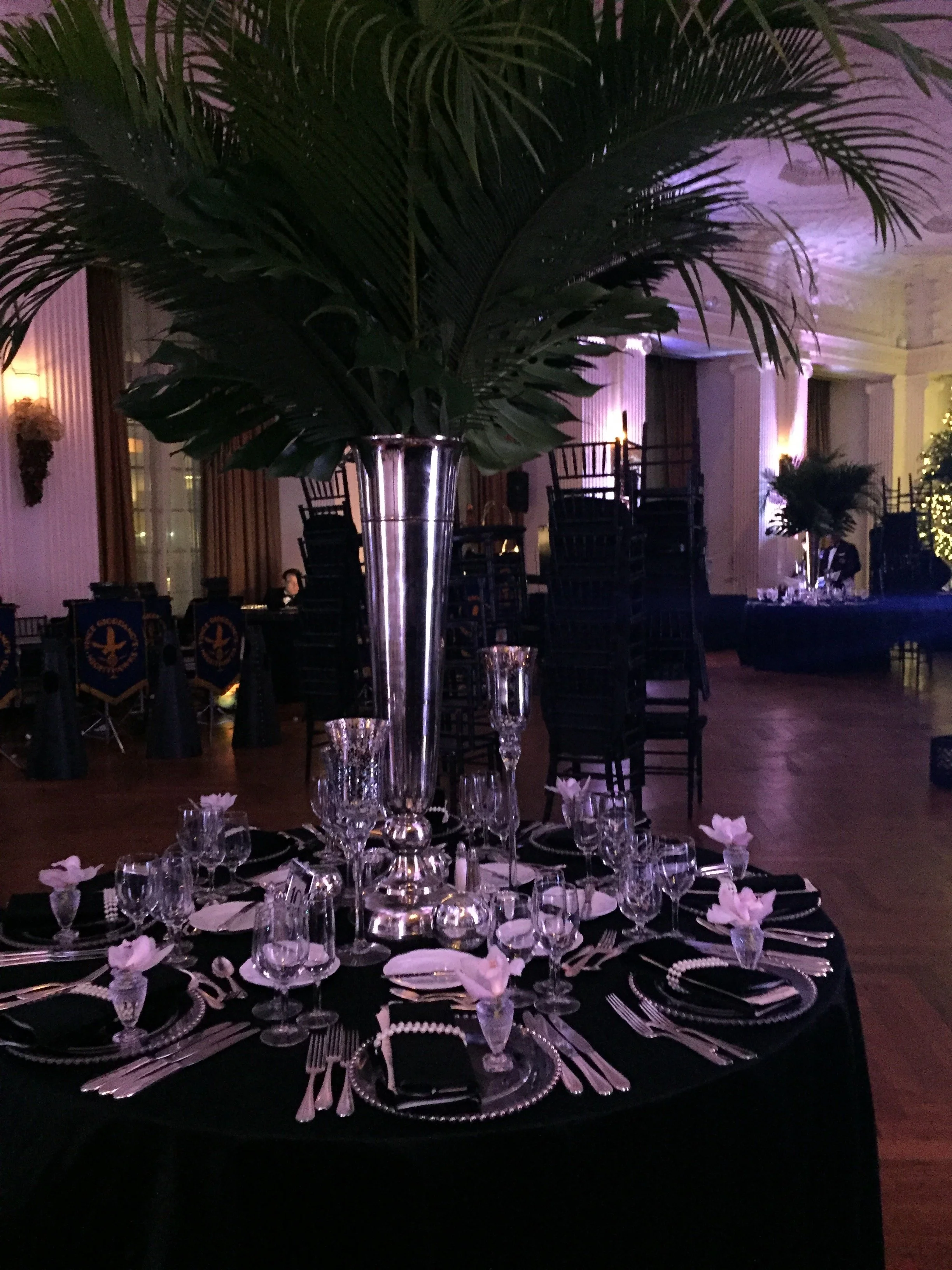 Elegant dinner table set with multiple wine glasses, small pink flower decorations, silverware, and a large centerpiece with palm leaves in a dimly lit banquet hall with purple uplighting.
