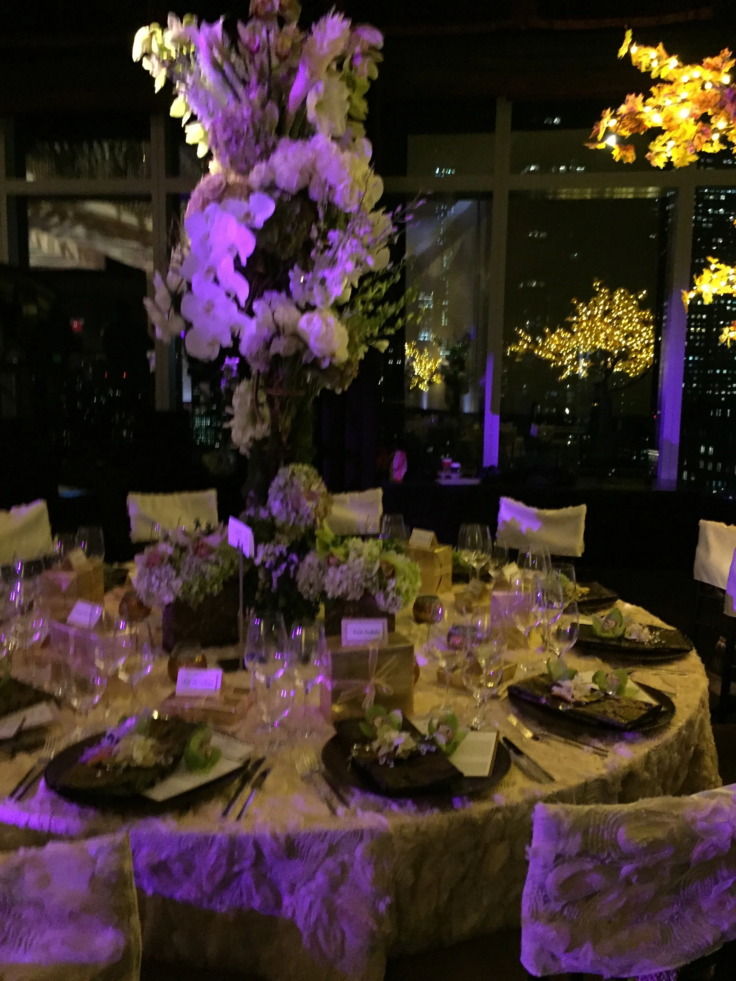 Elegant indoor event setup with a large floral centerpiece, table settings, and city skyline view through large windows at night, illuminated with purple and yellow lighting.