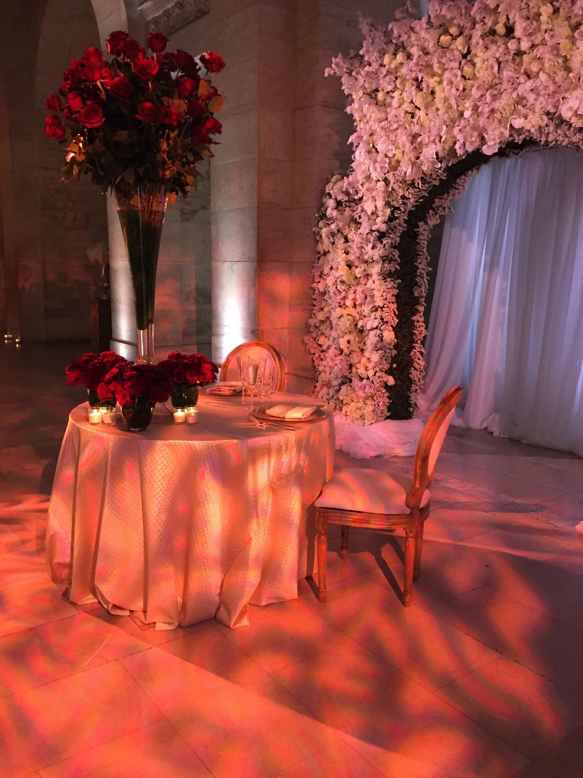 A decorated event table with a large bouquet of red roses in a tall glass vase, smaller red flower arrangements, and candles. In the background, there is a floral arch made of pink and white flowers with white curtains and a stone wall.