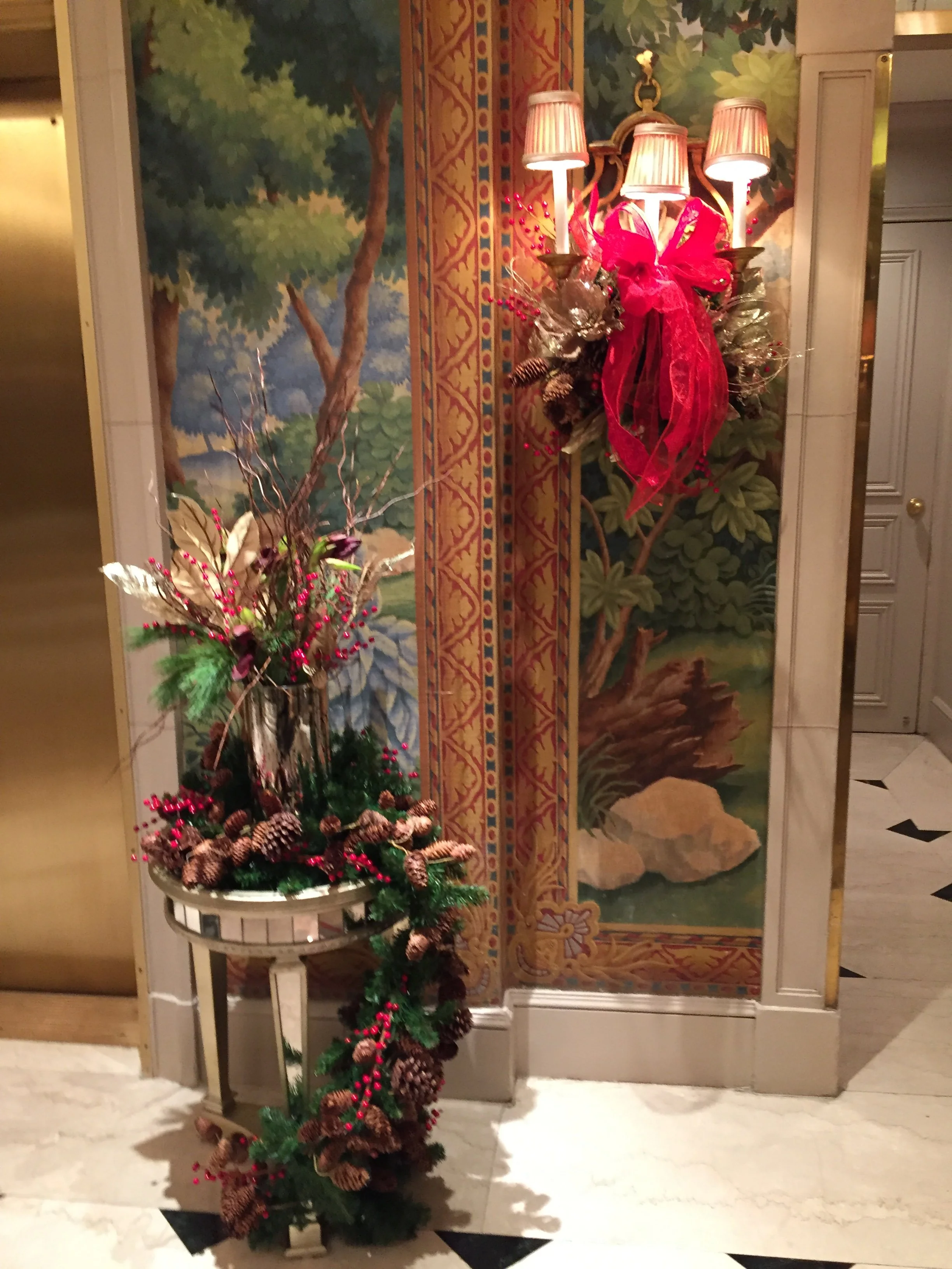 Decorative arrangement with pine cones, berries, and branches in a silver vase, positioned in front of a mural of trees and foliage, with a wall-mounted lamp fixture with three shades above.