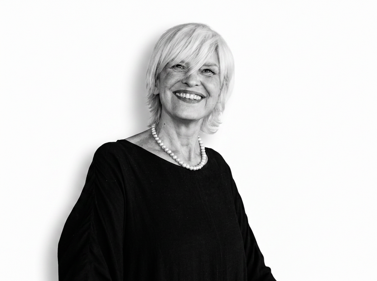 A smiling older woman with short, blonde hair, wearing a black top and a pearl necklace, standing against a plain white background.