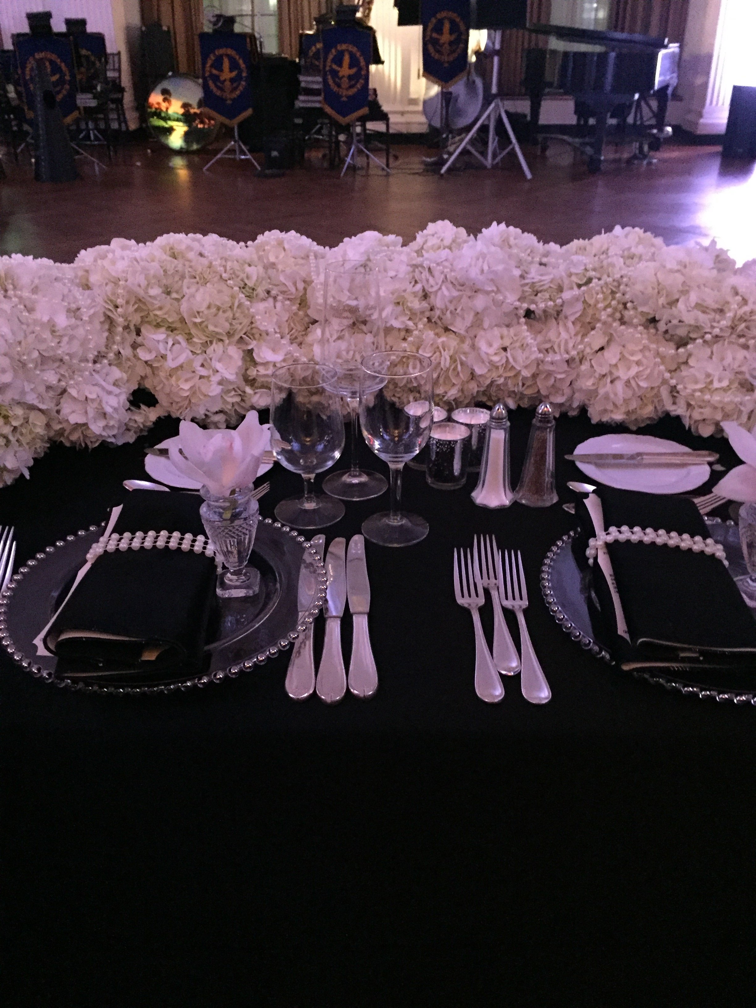 Table setting with black napkins, crystal glasses, silverware, and pearl necklaces, with a large floral centerpiece of white hydrangeas, in a dimly lit banquet hall with a stage and musical instruments in the background.