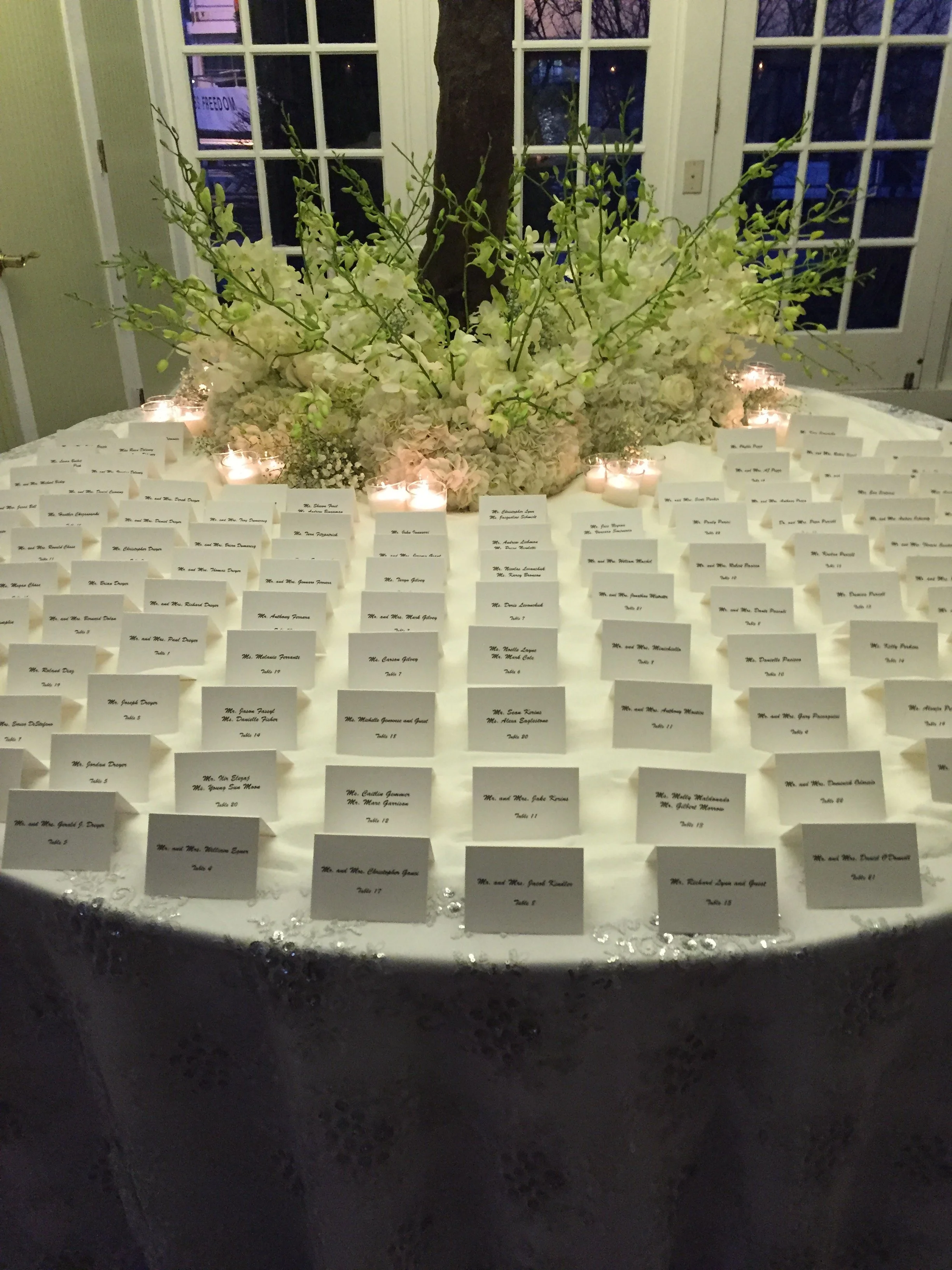 A table with a large floral centerpiece and numerous place cards, candles, and table numbers, likely for a wedding reception or formal event, set against a background of a door and windows.