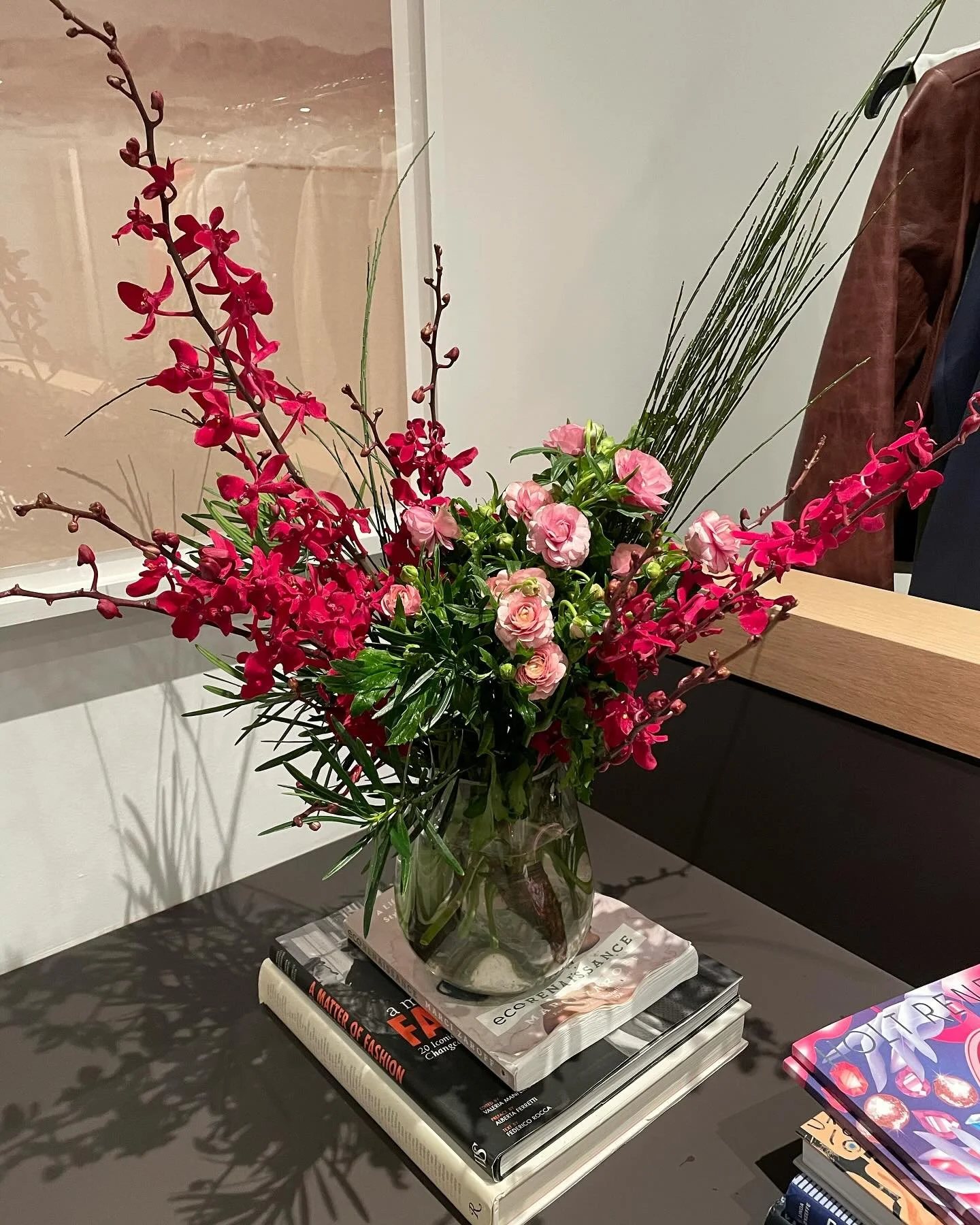 A glass vase filled with pink roses, red orchids, and tall green grasses, placed on a stack of books on a black table.