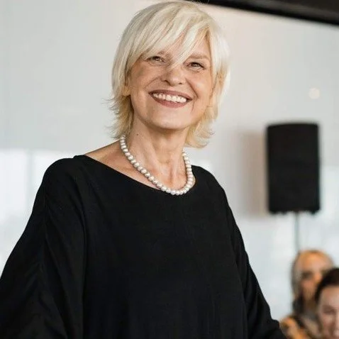 A smiling older woman with short white hair wearing a black top and pearl necklace, standing in an indoor setting with a speaker and several people in the background.