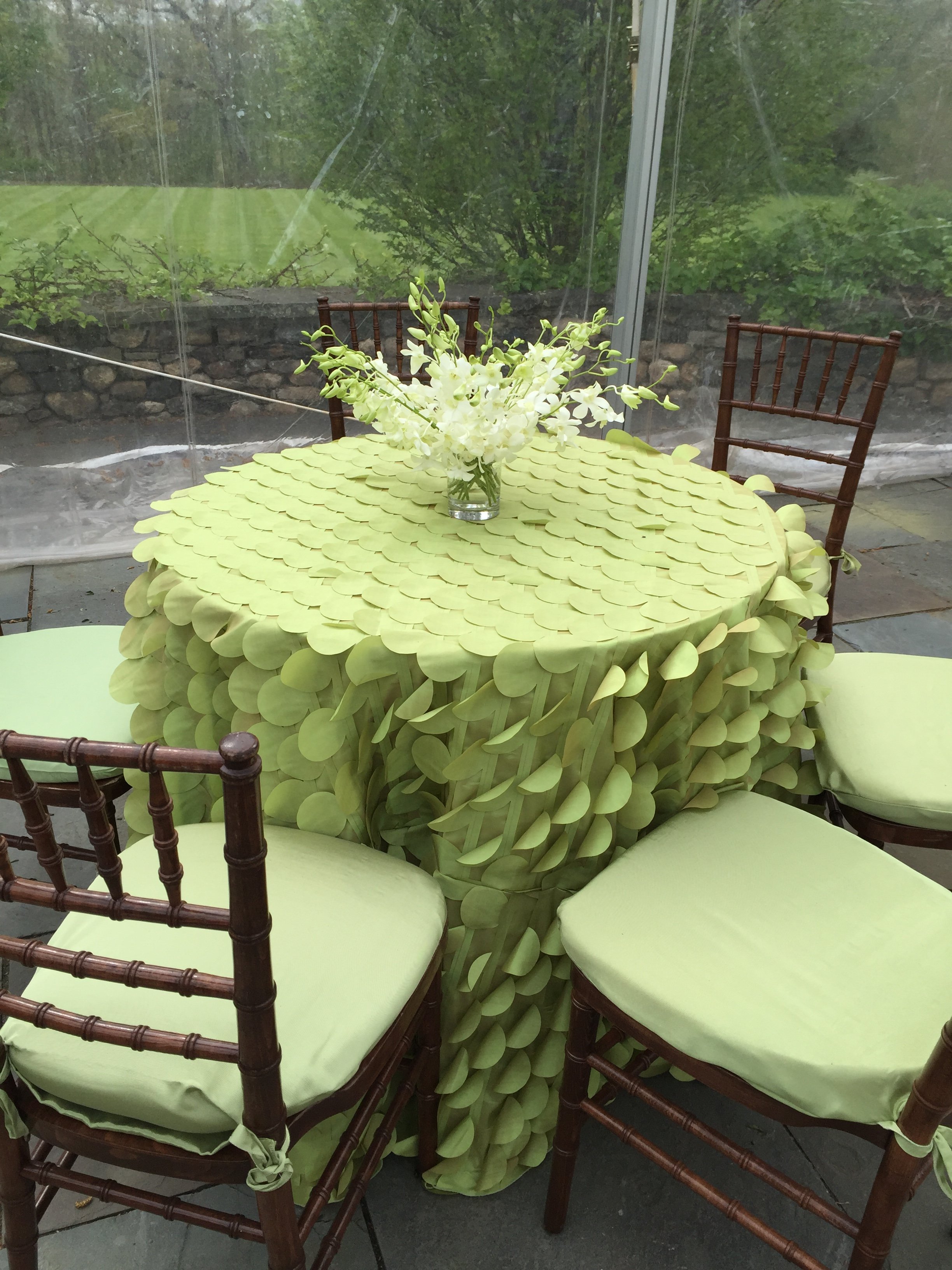 A round table with a pale green tablecloth, surrounded by four dark wooden chairs with light green cushions. On the table is a glass vase with white flowers. Behind the table is a large glass window showing a lush outdoor garden with green grass, bus