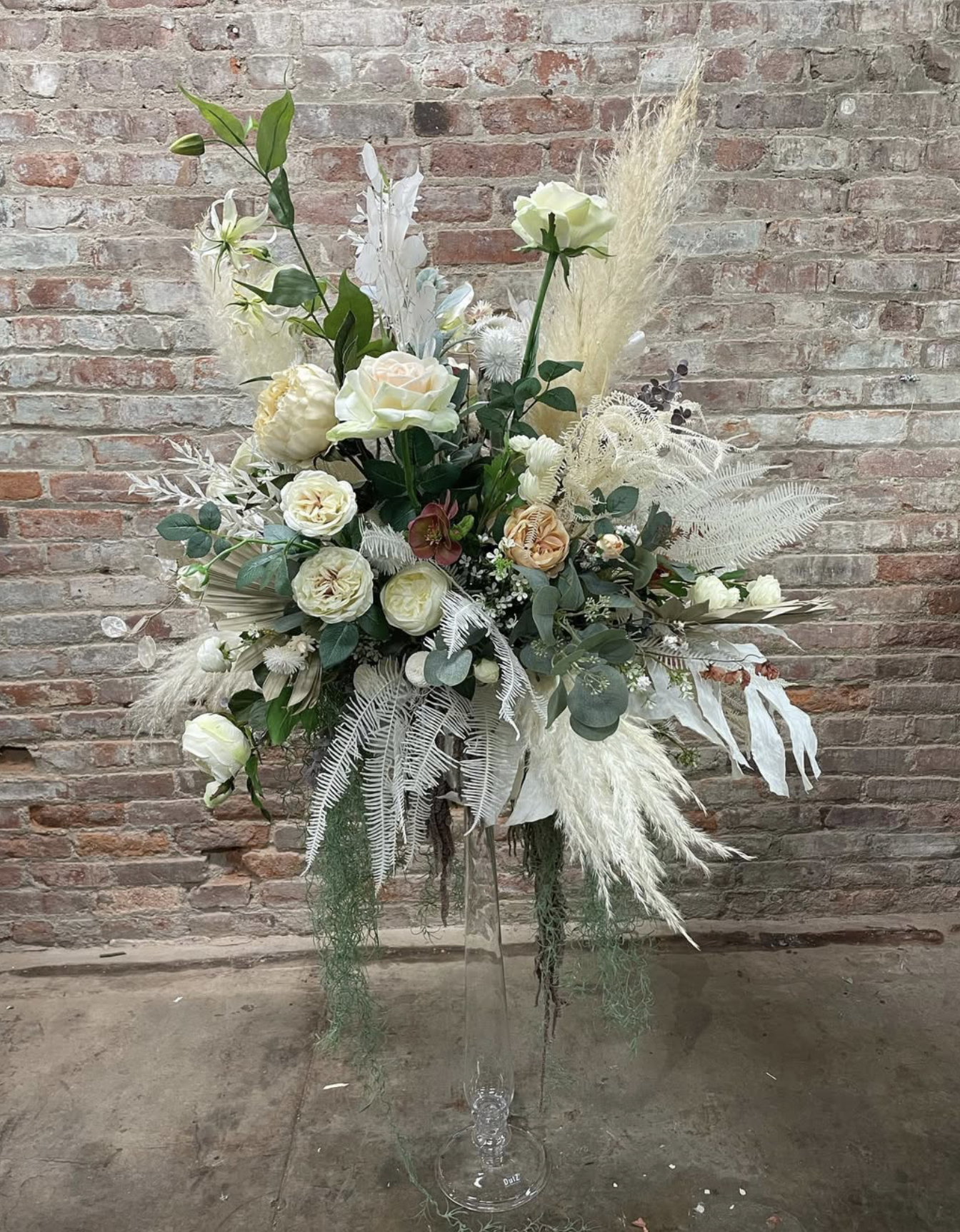 A flower arrangement with white roses, white lilies, white pampas grass, and greenery in a tall, clear glass vase against a brick wall background.