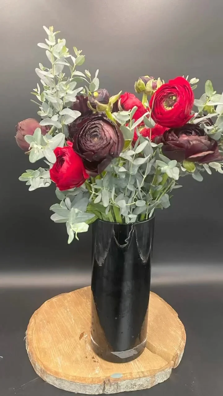 A black vase containing a bouquet of red, purple, and green flowers, placed on a round wooden slab, against a dark background.