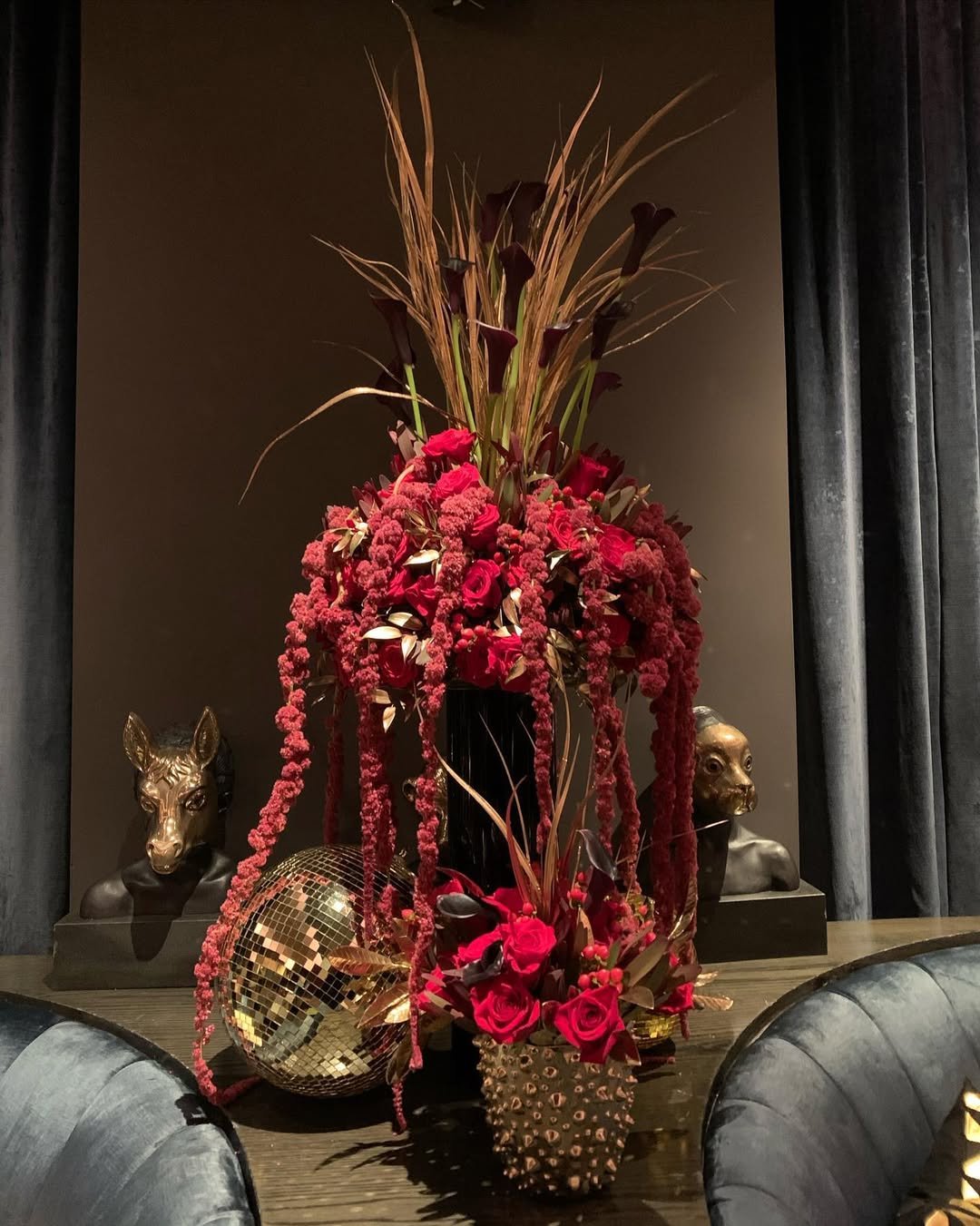 A tall floral arrangement with red roses, pink flowers, and dark leaves in a tall black vase, surrounded by gold animal busts and a gold disco ball, on a dark wooden table with dark curtains in the background.