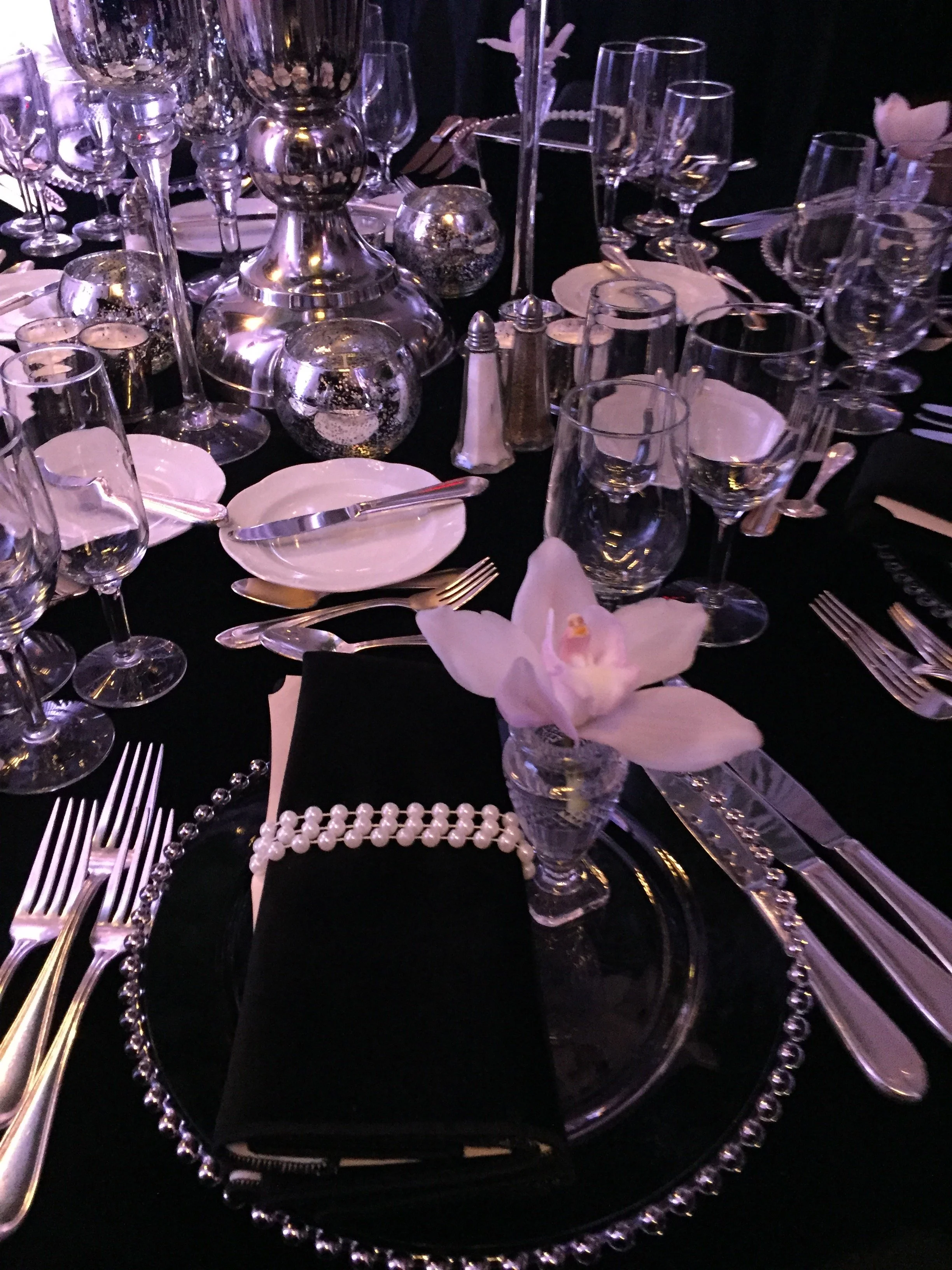A formal table setting with multiple wine glasses, water glasses, plates, silverware, a black napkin with a string of pearls, and a pink orchid in a glass vase, with decorative candles and silver vases on the table.