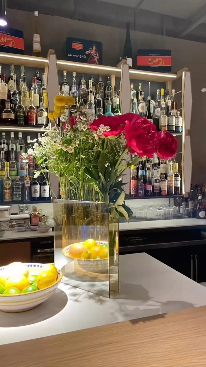 A bar countertop decorated with a large glass vase of pink and red flowers, and bowls of colorful fruit, with a backdrop of shelves filled with various bottles of alcohol.