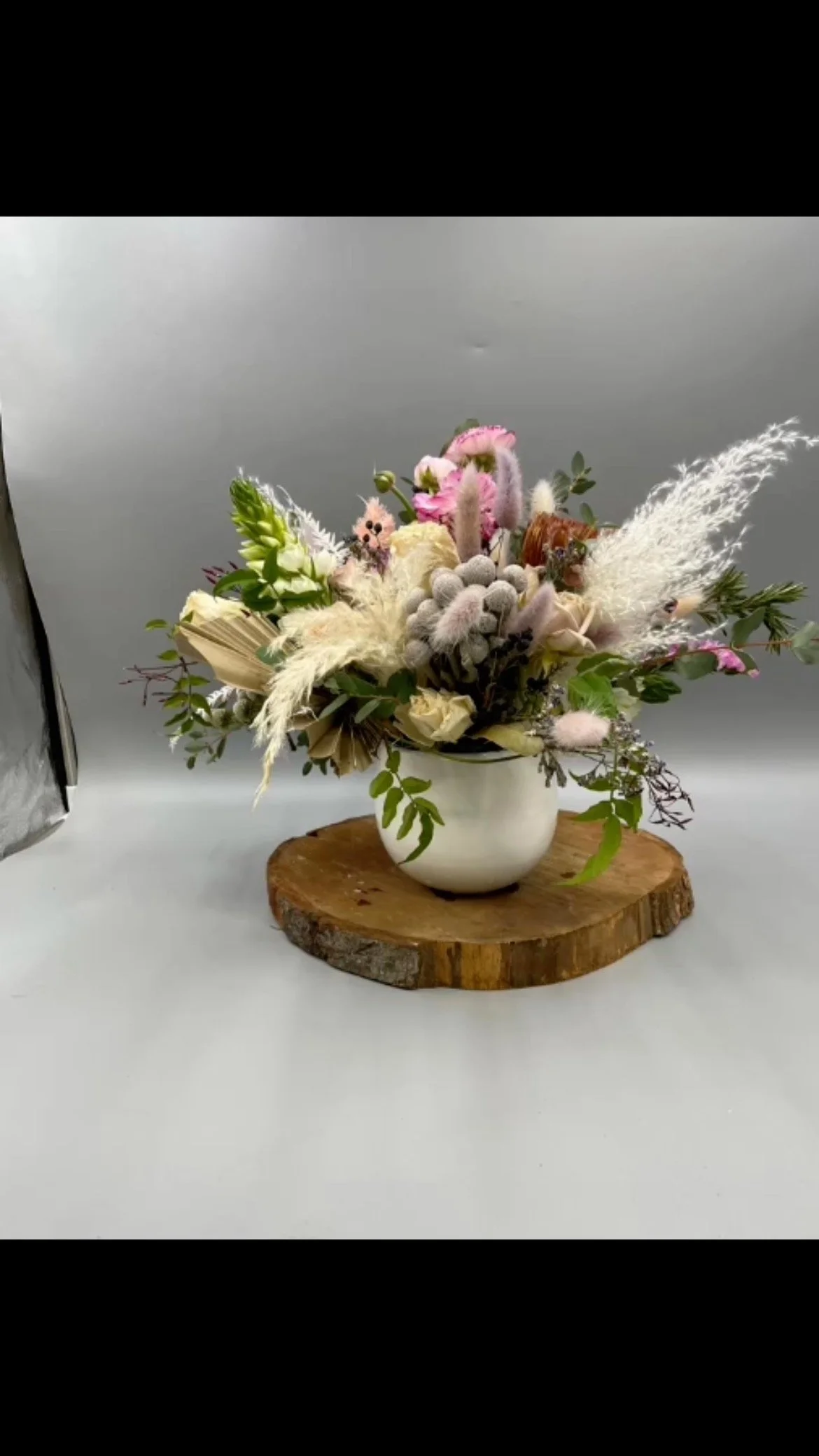 A bouquet of assorted flowers including roses, greenery, and dried flowers, arranged in a white vase on a wooden slab, with a plain background.