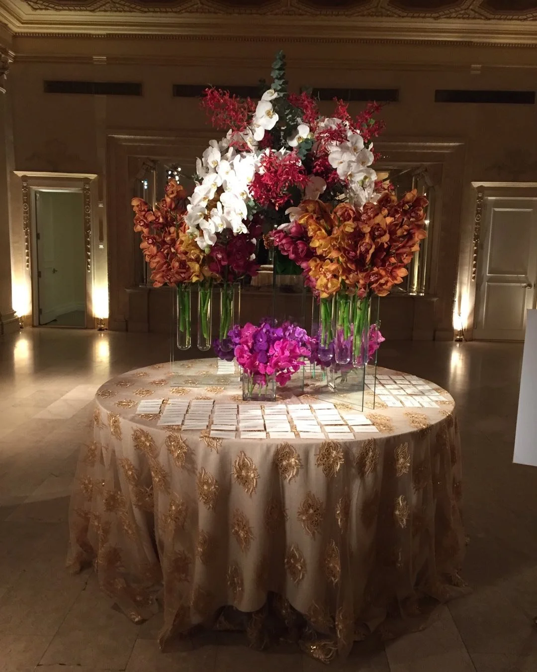 A round table with a gold embroidered tablecloth displays various flower arrangements in tall glass vases at an elegant event space.