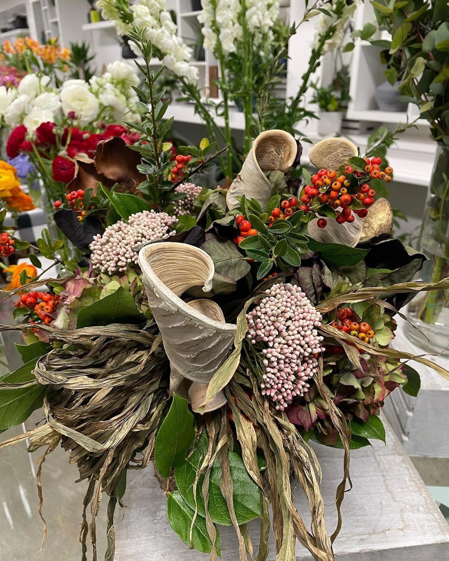 Arrangement of various flowers and foliage, including red berries, white dried flowers, and green leaves, with decorative dried and curled tree bark pieces.