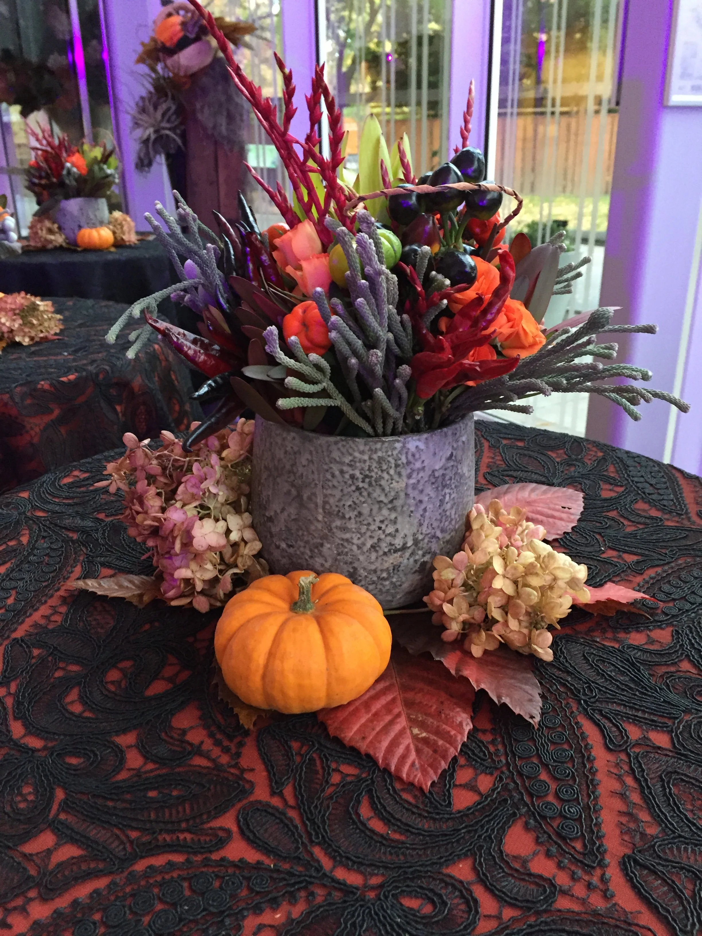 A pumpkin on a table surrounded by fall leaves and a floral arrangement in a vase, with a background of windows and trees.