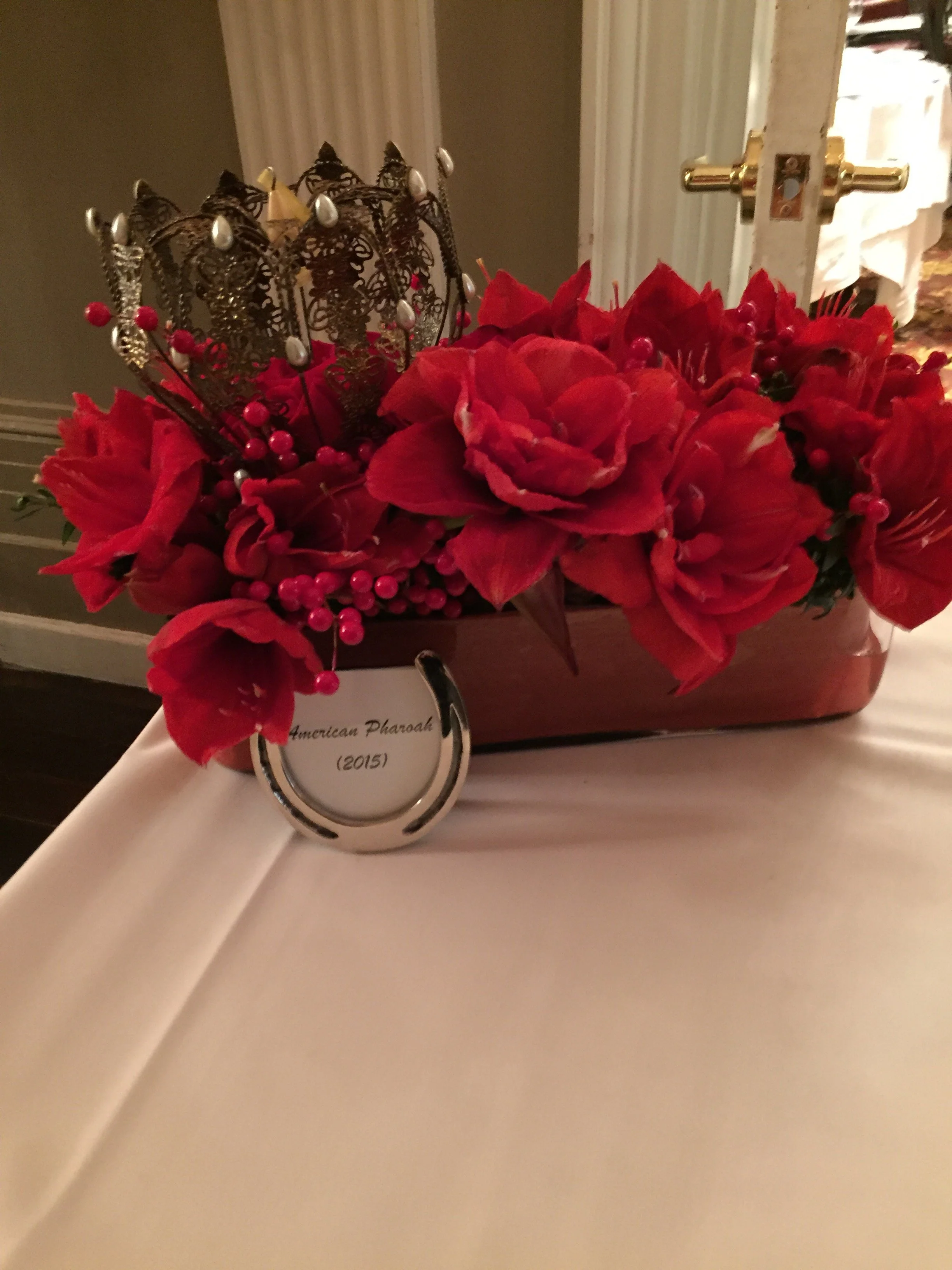 A decorative crown and a floral arrangement with red flowers and berries on a white surface, with a label reading "American Pharoah 2015" in front of the display.