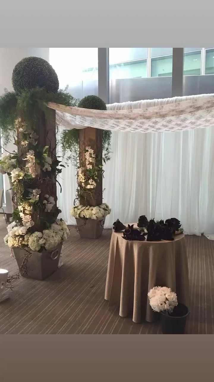 Floral decoration with two wooden posts draped with fabric and adorned with white flowers and greenery, with a small table holding dark floral arrangements and a potted white flower in front, set against white drapery and windows.