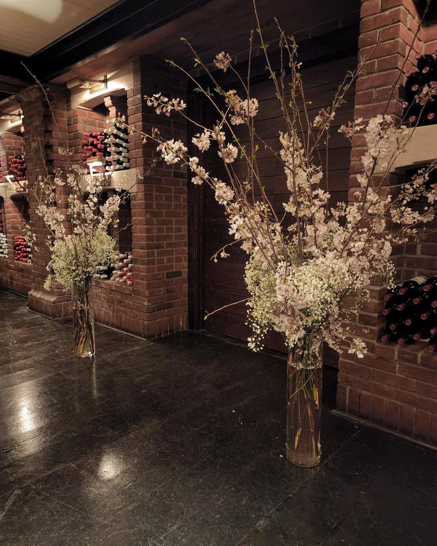 Two tall vases filled with branches of white flowers are placed on a dark floor in front of a brick wall with wine racks.