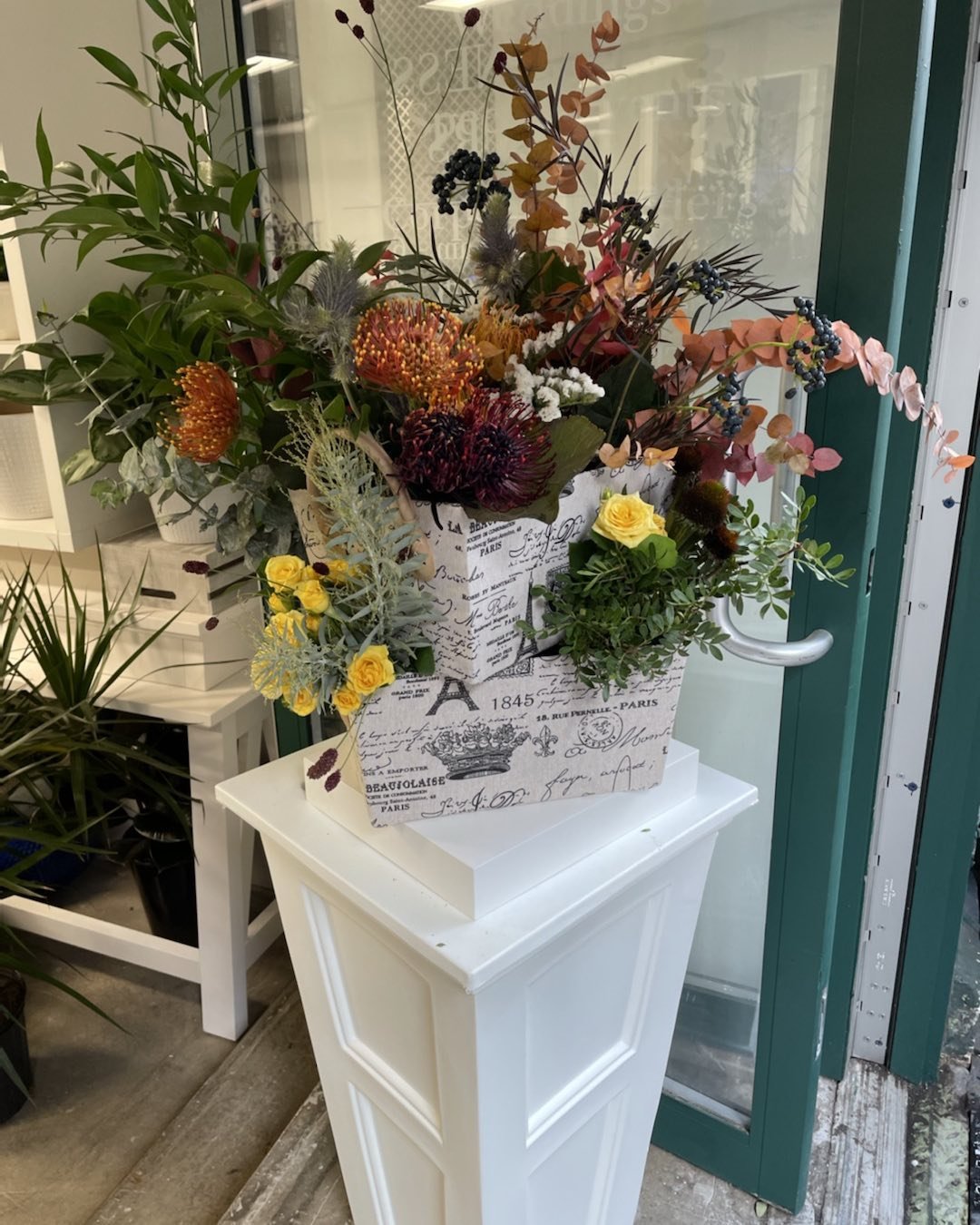 A floral arrangement in a decorative white box with vintage script, featuring a variety of flowers and greenery, displayed on a white pedestal near a glass door.