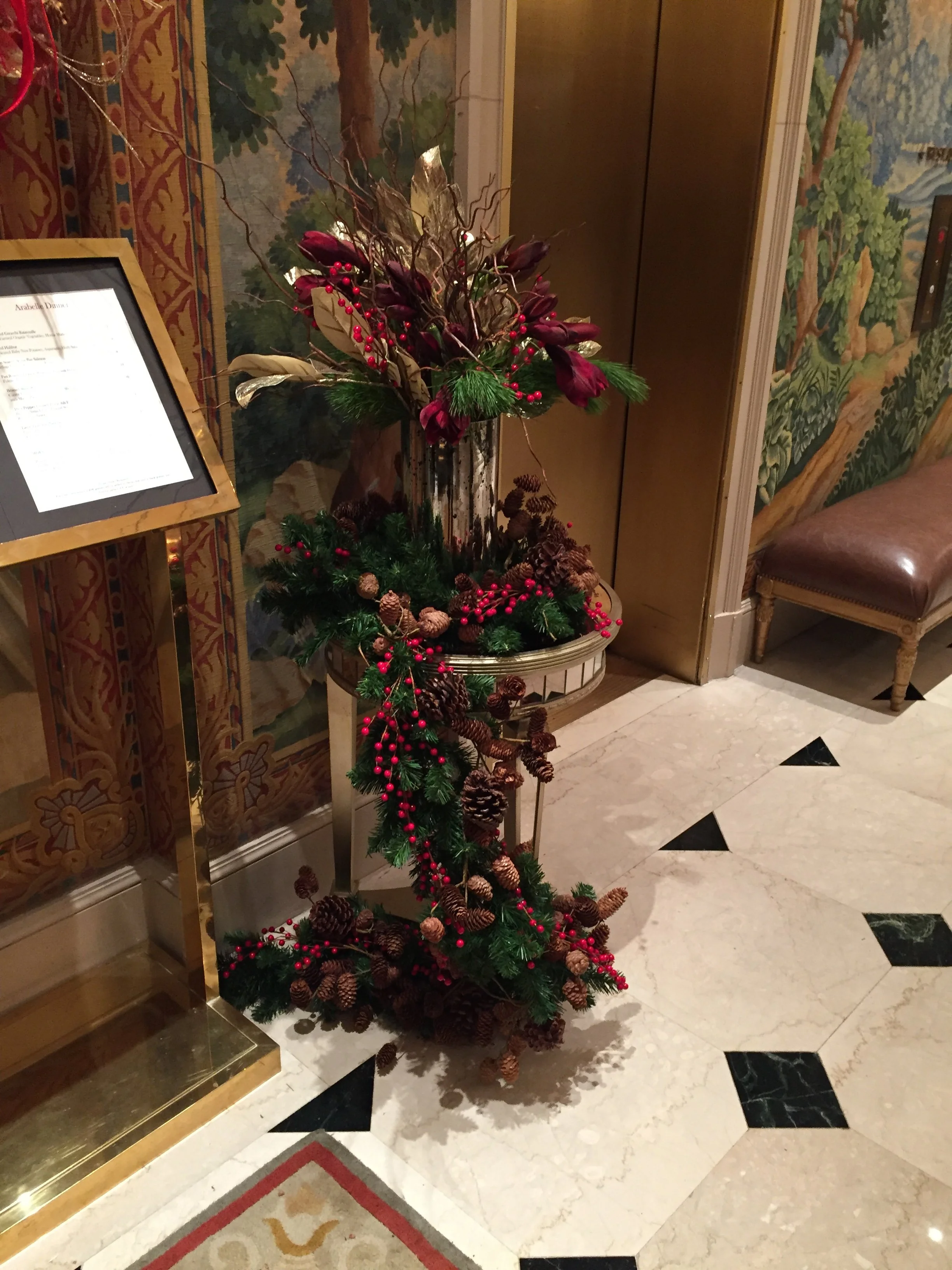 A Christmas-themed floral arrangement on a stand near an elevator, decorated with pinecones, red berries, green foliage, and dried branches inside a gold-trimmed pot in a hotel lobby.