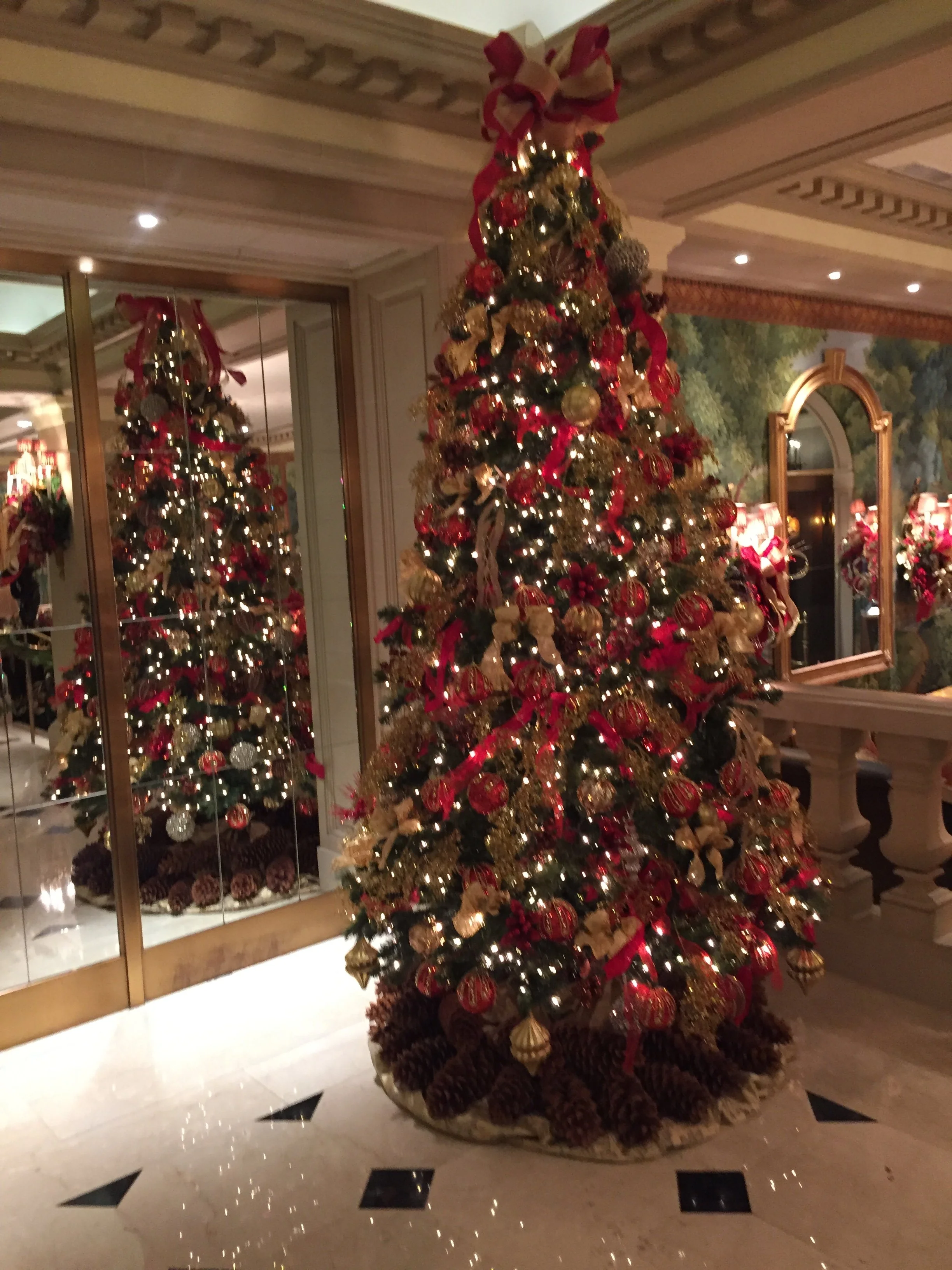 A decorated Christmas tree with red and gold ornaments, ribbons, and lights, placed indoors near a mirror and a wall with a mural. Pinecones are arranged at the base of the tree.