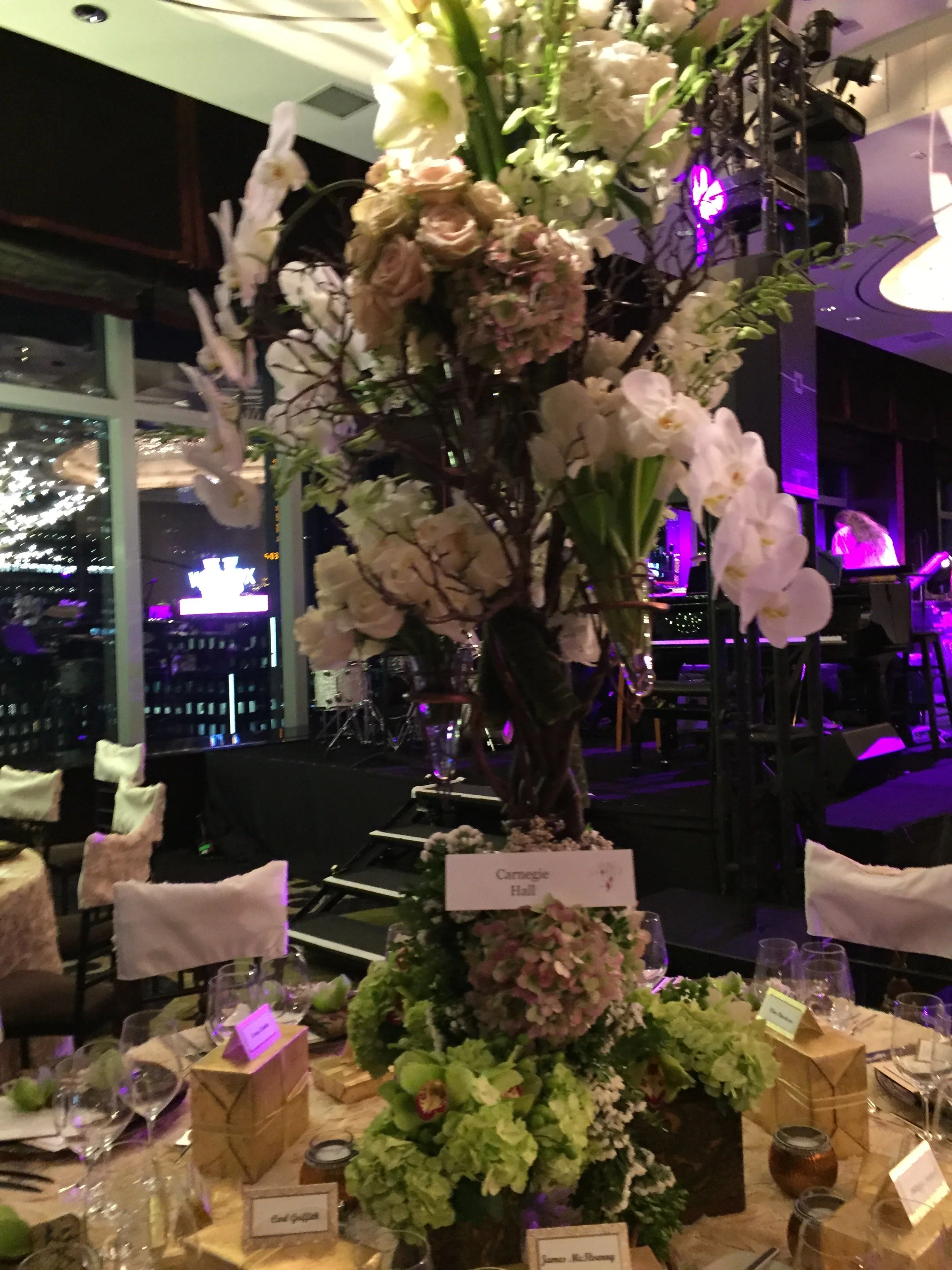 A floral centerpiece with large white and pink roses, hydrangeas, and various greenery at a banquet table with wine glasses and place cards, in a dimly lit event venue.