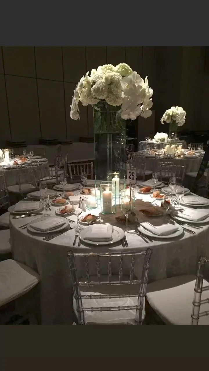 Elegant banquet table with a tall floral centerpiece of white flowers, white napkins, glassware, candles, and place settings for a formal event, dimly lit
