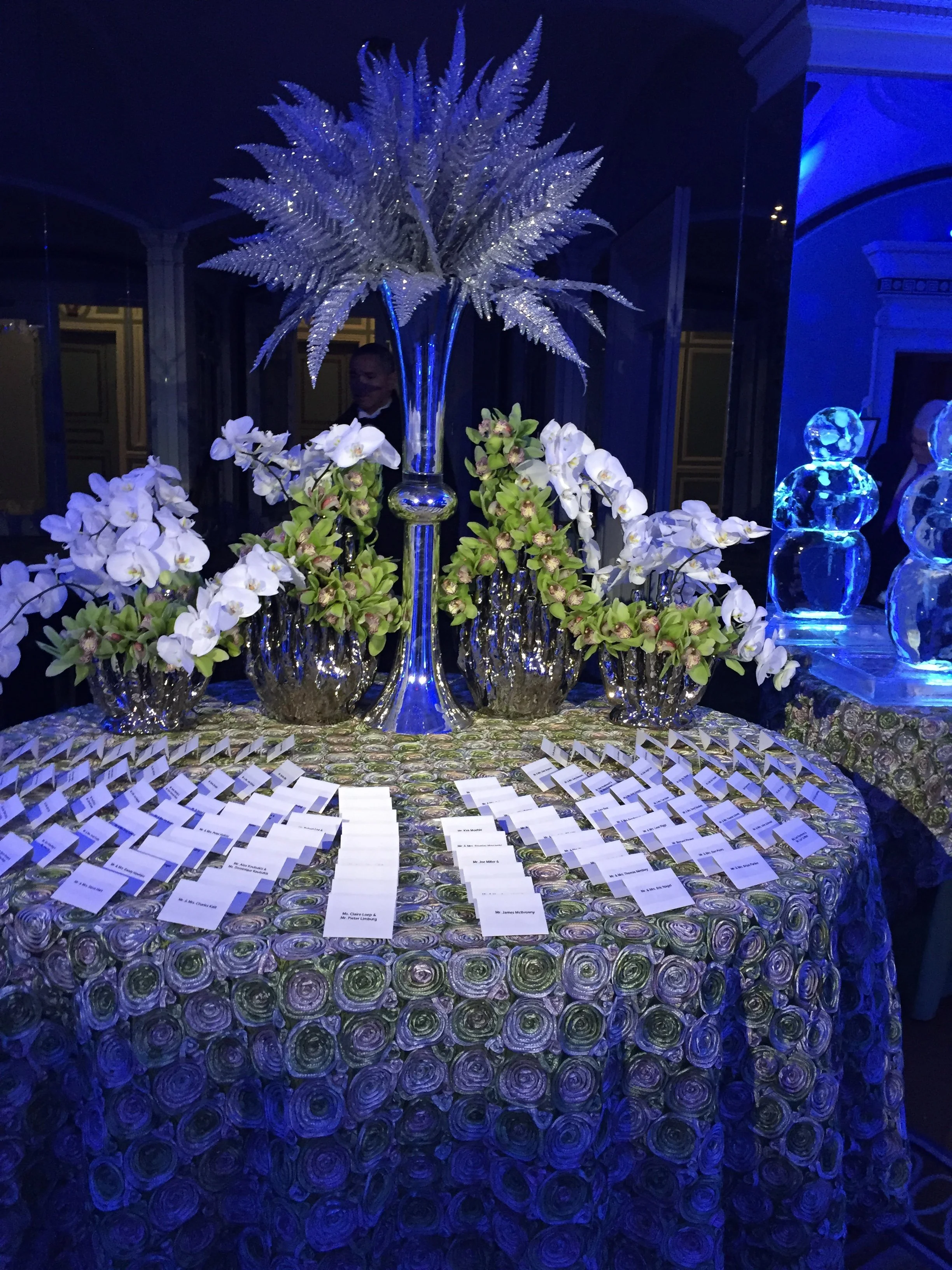A large round table decorated with an elaborate flower arrangement featuring tall, crystal-stemmed vase with a large arrangement of sparkling purple leaves, surrounded by white orchids and greenery in crystal vases, with numerous place cards laid out