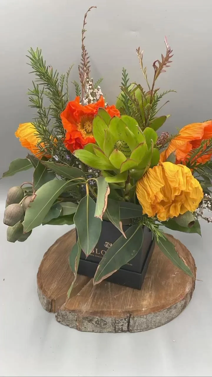 A floral arrangement with yellow and orange flowers, green foliage, and seed pods in a black square container on a wooden slab.