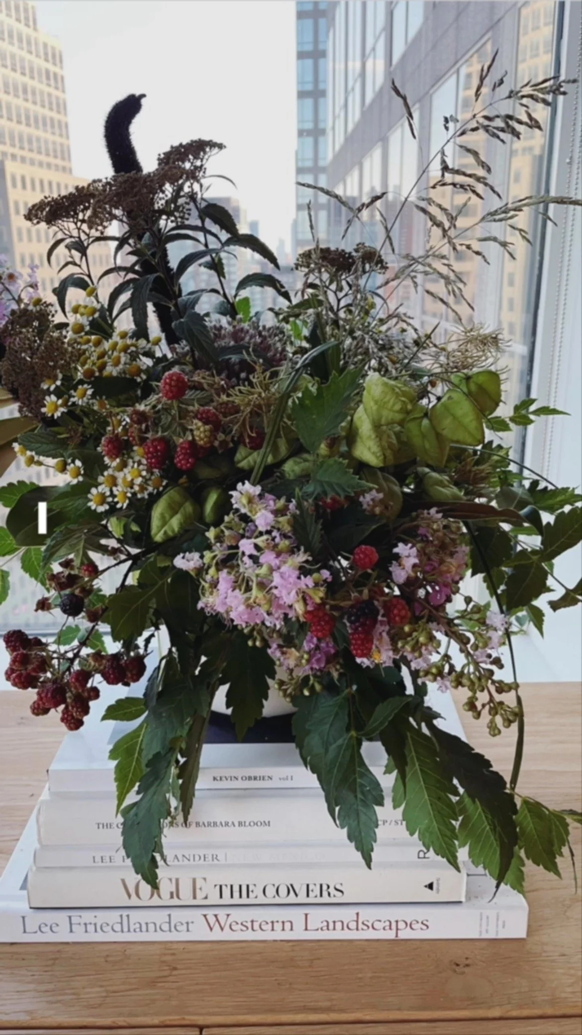 A bouquet of mixed flowers and foliage arranged on top of a stack of large art books, with city buildings visible through a window in the background.