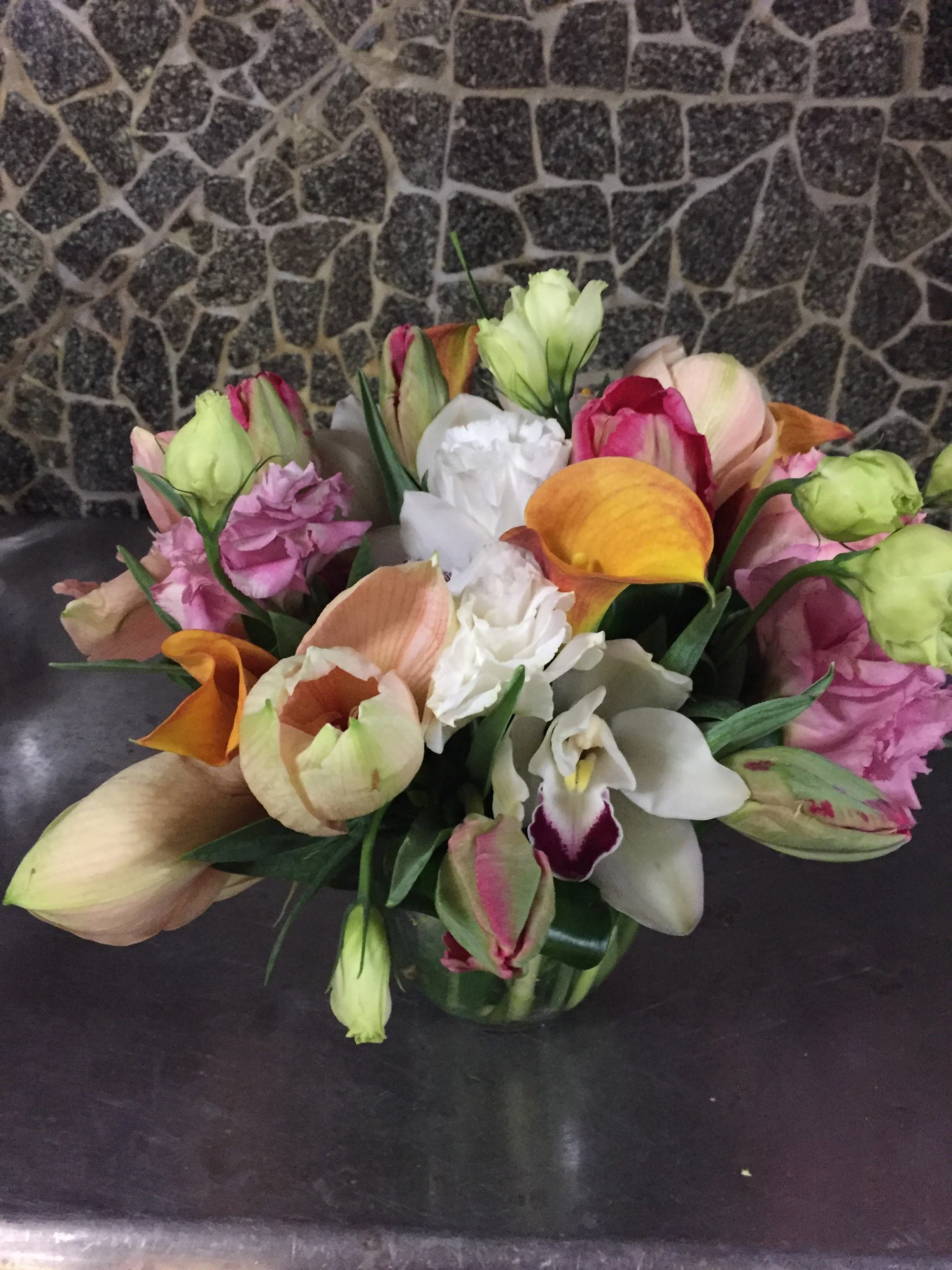 A floral arrangement with various flowers including roses, orchids, and tulips in a glass vase, placed on a dark surface with a textured stone wall in the background.