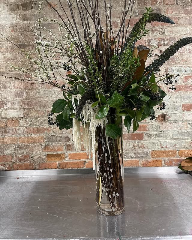 A tall glass vase filled with mixed greenery and decorative branches, placed against a brick wall.